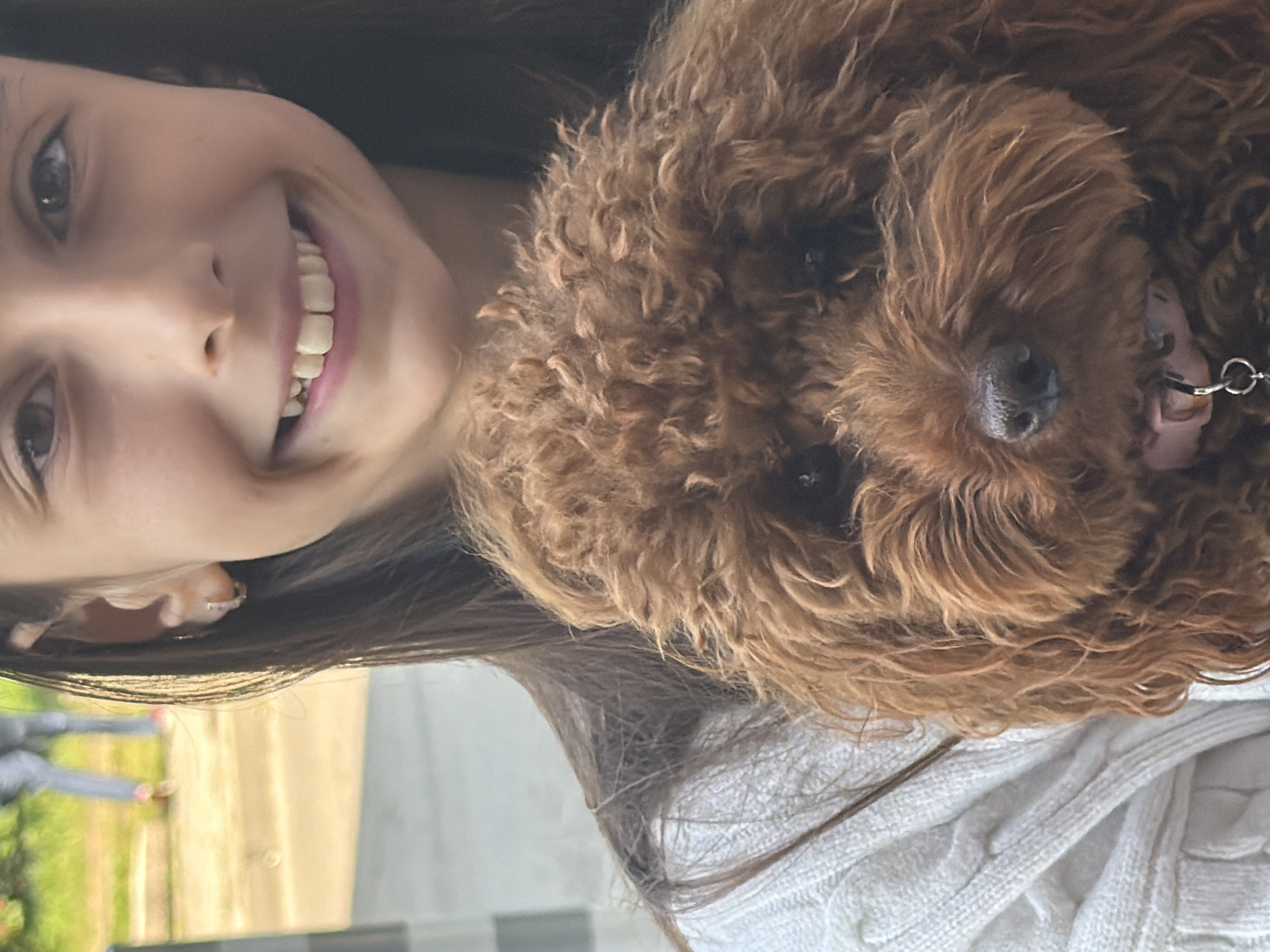 The image depicts a young girl holding a reddish-brown curly-haired dog outdoors, with the scene located in Sofia, Bulgaria. The girl, positioned in the upper right and center of the frame, has long dark hair, brown eyes, and is smiling directly at the camera, revealing her upper teeth. She wears a white cable-knit sweater and a small, light-colored hoop earring is visible in her left ear. The dog, occupying the lower central portion, features dense, curly reddish-brown fur, dark eyes, and a dark nose. A pink collar with a silver tag is visible around its neck. The background is heavily blurred, showing green foliage on the left, a light-colored ground surface, and the lower body of a distant individual wearing blue jeans and red shoes. The scene is illuminated by natural daylight.