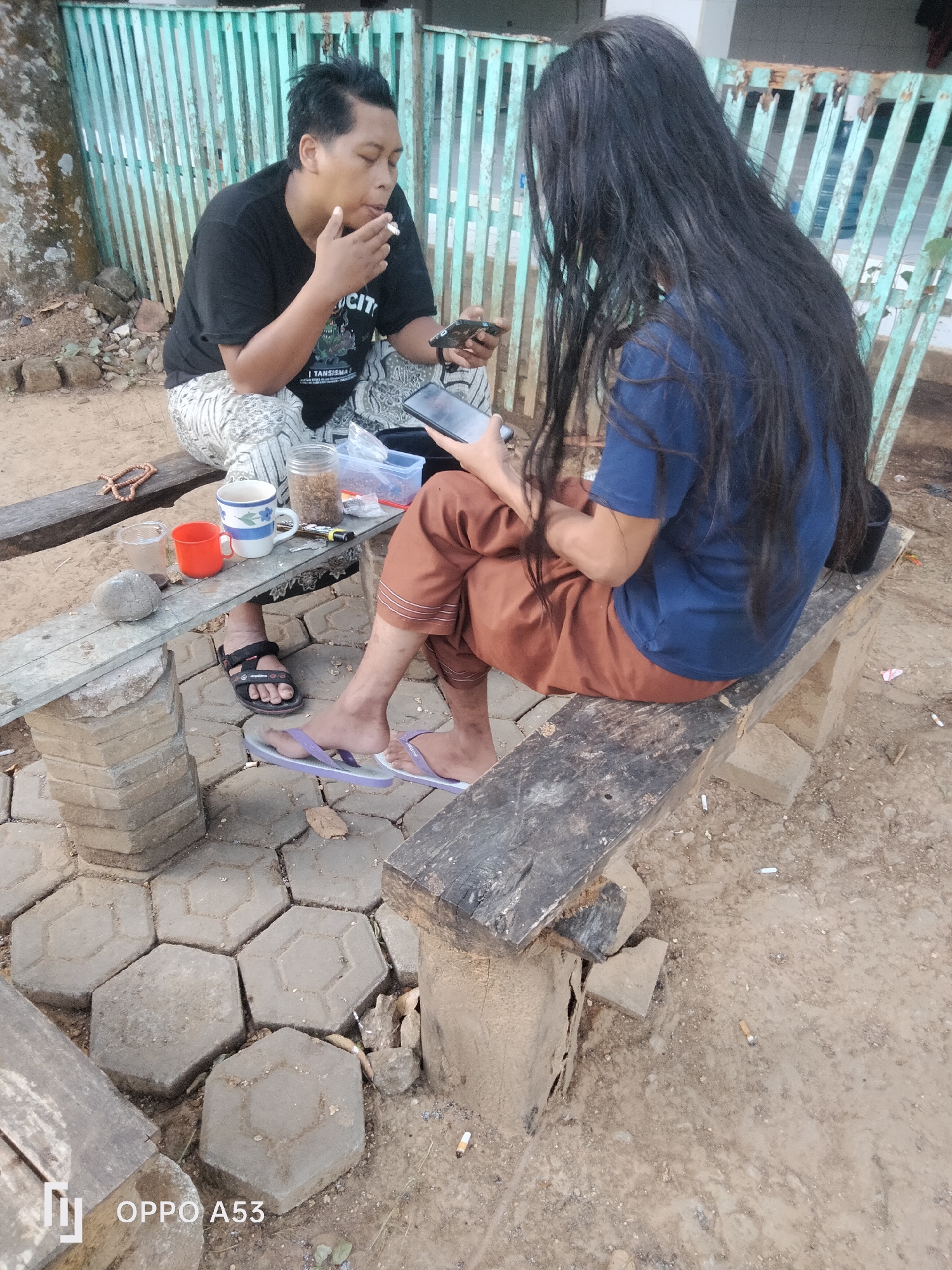 In Badung, Indonesia, two individuals are seated on rustic wooden benches outdoors. The person on the left, with short black hair, is smoking a cigarette and holding a smartphone. They are wearing a black t-shirt and patterned pants. Next to them are a red cup, a white mug with blue floral designs, a small glass, a stone, and a jar containing what appears to be tobacco. A beaded bracelet is also visible.
The person on the right has long, dark hair and is wearing a blue t-shirt and brown pants. They are also holding a smartphone. Both individuals are barefoot or wearing sandals. The ground is paved with hexagonal stone tiles and dirt. A faded turquoise metal fence is in the background, and discarded cigarette butts are scattered on the ground. The scene suggests a casual outdoor gathering.