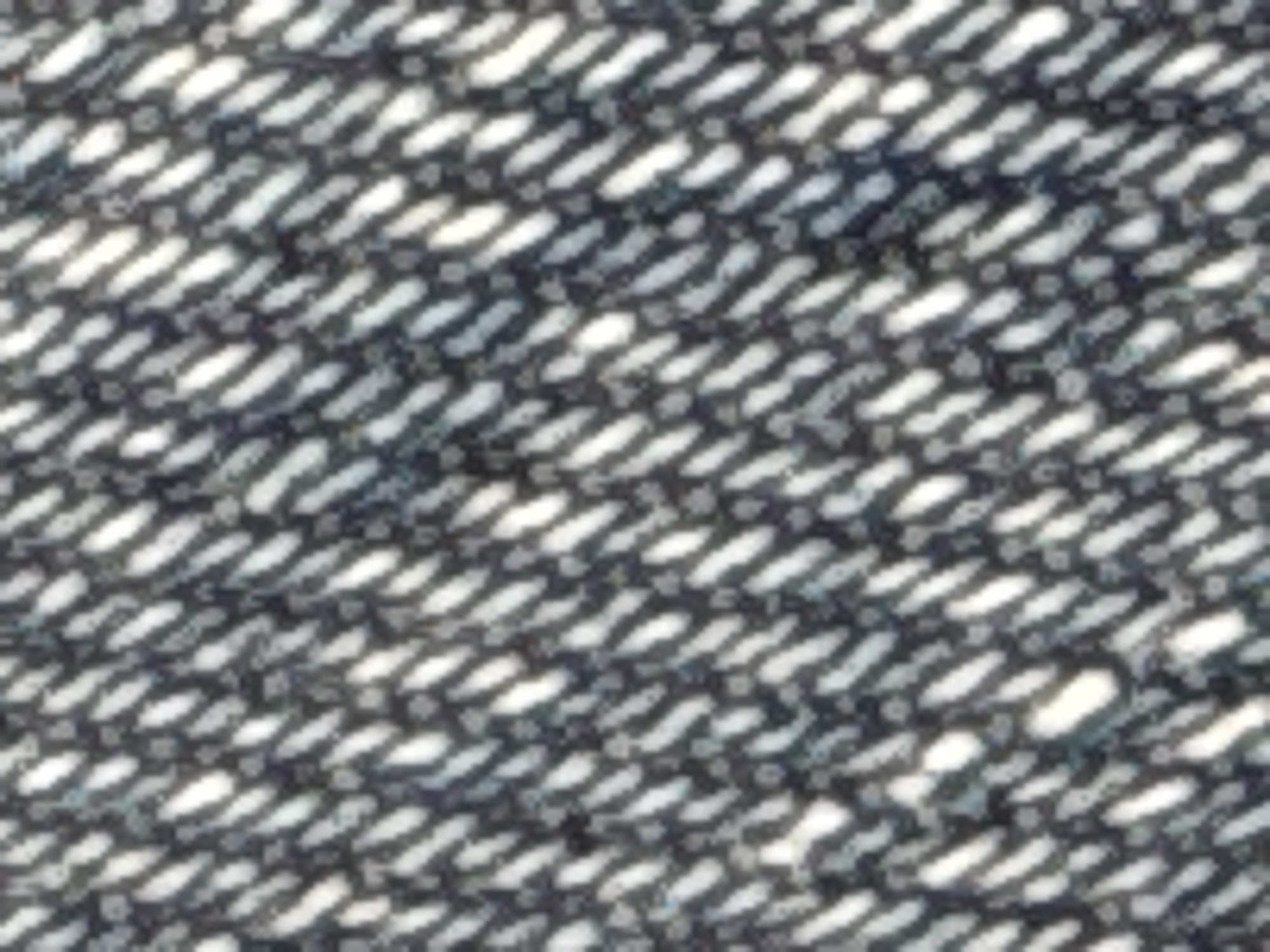 The image is a close-up, abstract view of a dark, textured fabric with a diagonal weave pattern. White or light-colored streaks and small, circular dots are interspersed within the dark threads, creating a speckled or dappled effect. The lighting appears to be uneven, with some areas being brighter and others darker, suggesting shadows or highlights on the material. The texture is rough and appears to be woven, possibly denim or a similar coarse fabric. There are no discernable people, objects, actions, interactions, or environmental features that would place this image in a specific city or location.