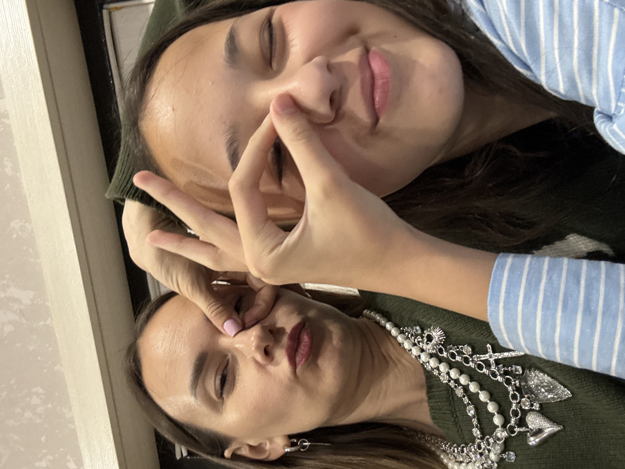 Two females pose closely together. The person on top has her eyes closed, a hand near her nose, and wears a blue and white striped shirt. The person below has one eye slightly open, a hand near her eye, and wears a green shirt with a pearl and silver necklace. Both have brown hair. The location may be Sochi, Russia.