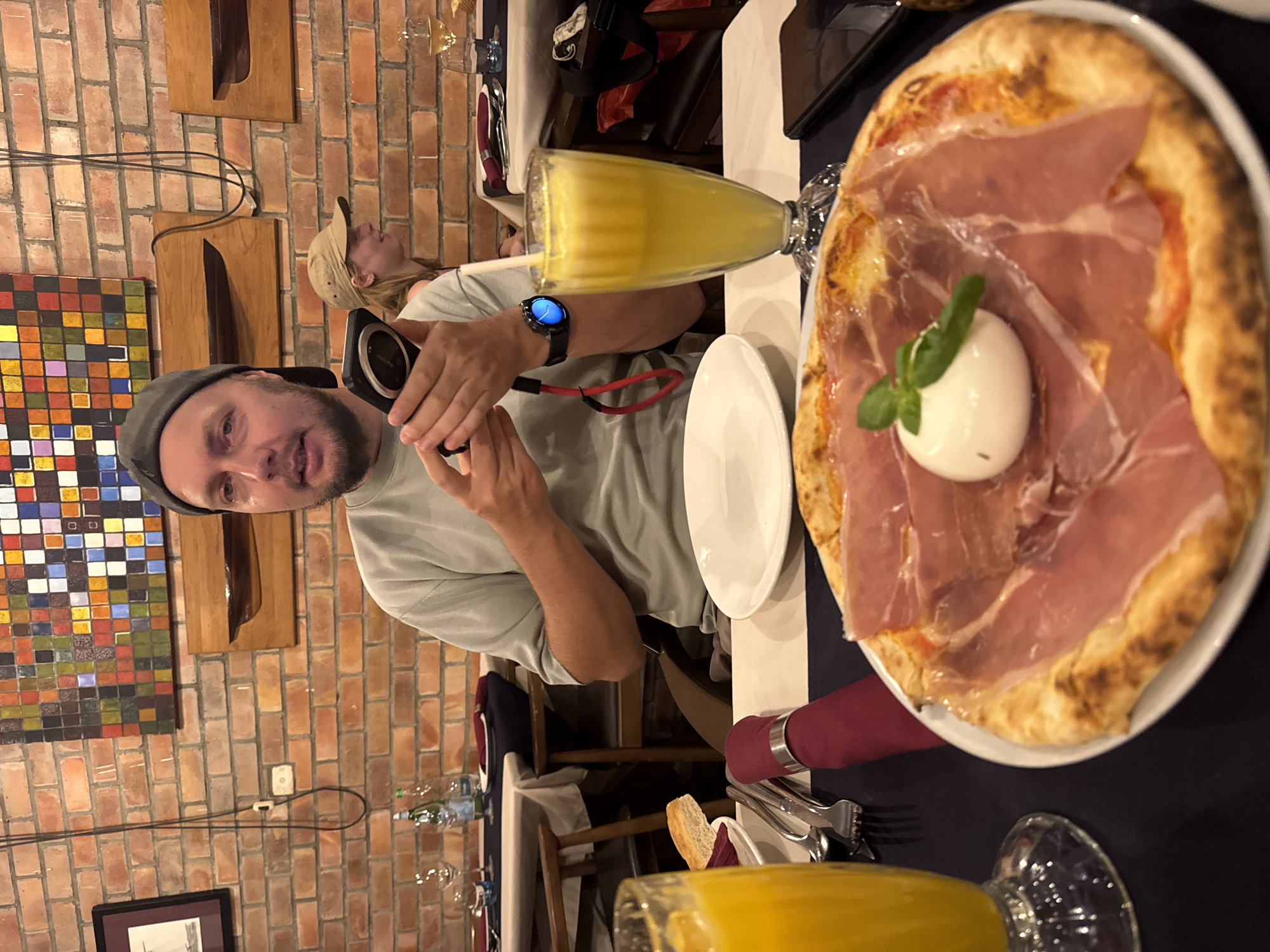 A man in a gray cap is taking a photo at a restaurant in Hà Nội, Vietnam. In the foreground, there is a pizza with prosciutto and burrata, along with a glass of orange juice. He is sitting at a table with another person in the background. The restaurant has a brick wall decorated with a colorful artwork and wooden knife displays. Other details include silverware, a bread slice, a water bottle, and additional glasses on the table.