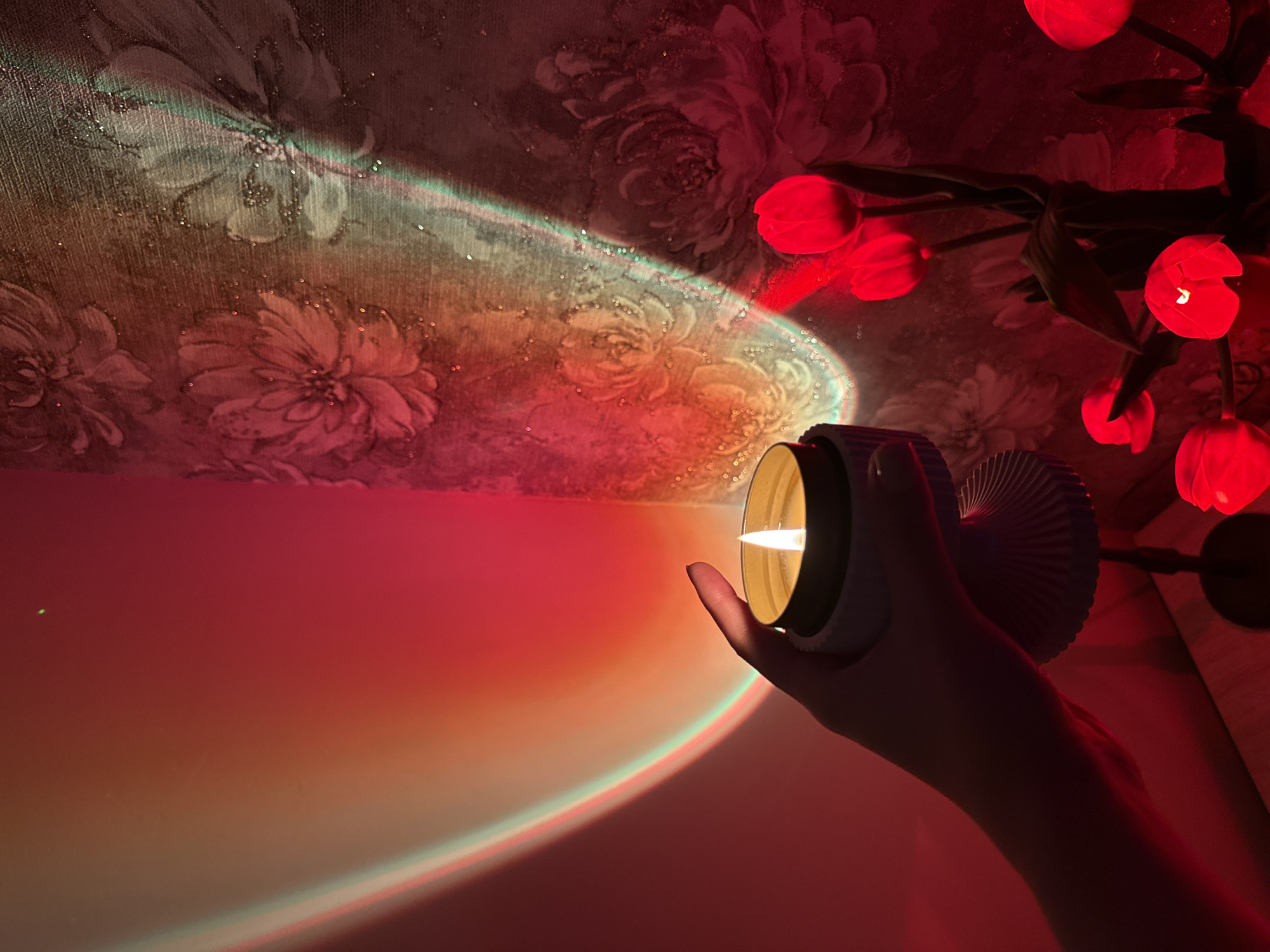 A hand holds a lit candle in a room illuminated by a sunset lamp and red tulip lights. The sunset lamp casts a gradient of orange, red, and green light onto the wall and textured wallpaper with a floral pattern. The lit candle has a bright yellow flame and is in a pale blue holder. In the foreground, red tulip lights cast a warm glow on a dark surface. The scene appears to be an indoor setting, possibly a home or studio.
