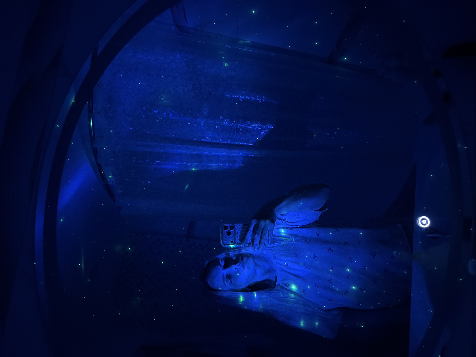 A person is depicted lying on their back in a dark, enclosed space, illuminated primarily by a vibrant blue light. Numerous small, glowing green dots are scattered across the surfaces, creating an immersive "starry night" effect. The individual, who appears to be a woman, occupies the lower central portion of the frame, looking upwards. She holds a smartphone with a clear case and a visible triple-camera system above her head with her right hand, suggesting she is taking a selfie or recording her surroundings. Her expression is neutral, and she wears a light-colored top patterned with repeating symbols that faintly glow under the blue light.
The setting features dark, possibly curved walls and ceilings, contributing to a capsule-like or intimate environment. Structural elements, including a dark, curved band with metallic details, are visible in the upper left. A distinct circular light fixture with a bright white ring is visible on the right side of the frame against the dark wall. The predominant blue and green lighting establishes an ethereal, futuristic, or serene sensory atmosphere, indicative of a relaxation room, a specialized spa, or an art installation. The overall mood is calm and contemplative. This scene is located in Votkinsk, Russia. No discernible text is visible in the image.