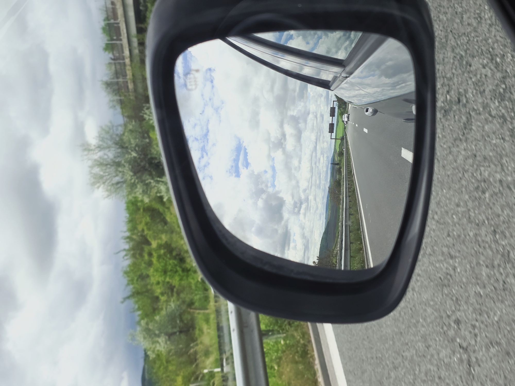 A car side mirror reflects a highway with a passing vehicle and grassy hills under a cloudy sky. The location is likely near Nevsha, Bulgaria, given the context. The mirror's frame is visible in the foreground, and a guardrail is seen alongside the road.