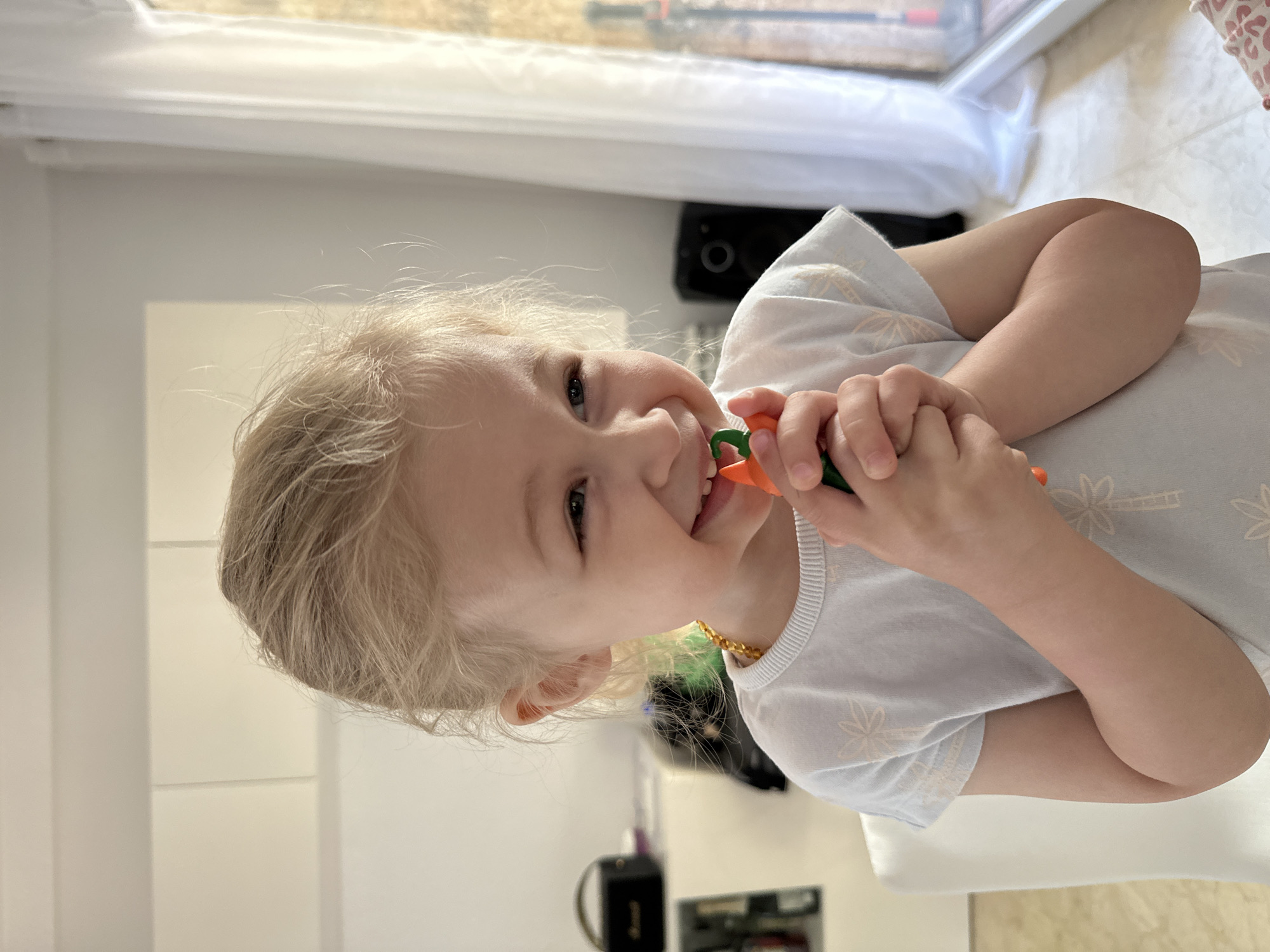 In a brightly lit indoor setting, a young Caucasian girl with blonde hair is captured from the chest up. She is seated in a white chair and is wearing a light blue t-shirt with a palm tree pattern and an amber necklace. The girl is holding a small orange and green object, possibly a toy or a crayon, up to her mouth and is smiling broadly. Her eyes are directed to her right, and her face is tilted slightly upwards. In the background, a white cabinet is visible to the left, and sheer white curtains hang to the right, with a window beyond them. The overall scene suggests a casual, home environment.