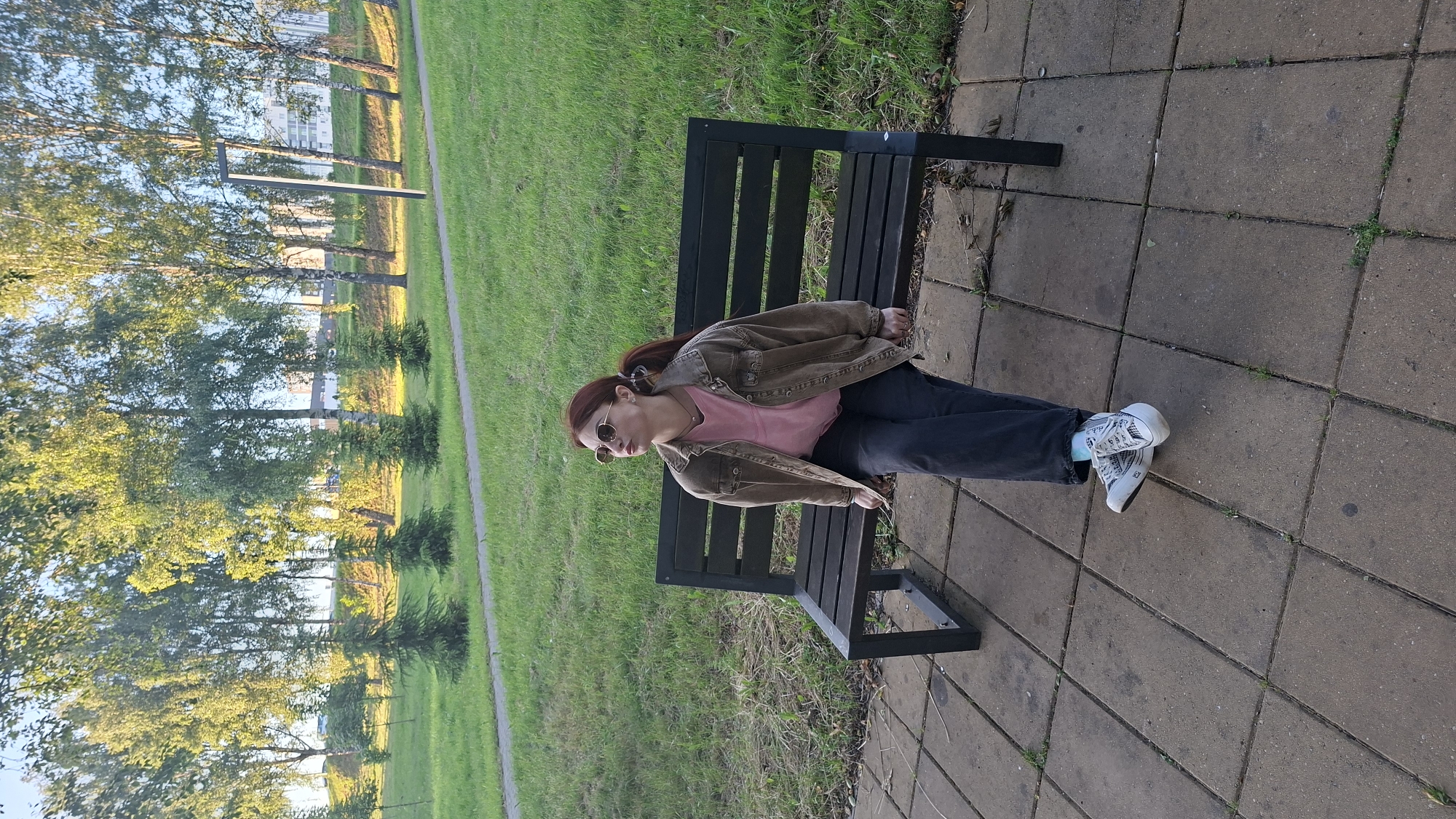 A young woman with auburn hair tied in a ponytail sits on a park bench in a city park in Smaliavicy, Belarus. She is wearing a light pink t-shirt, a dark brown denim jacket, dark grey jeans, white sneakers with black detailing, and round sunglasses. The bench is made of dark wood slats and black metal supports, positioned on a paved area of rectangular stone tiles. Behind the bench, a grassy area slopes upwards, lined with mature birch trees and smaller evergreen trees. In the background, several modern apartment buildings are visible, suggesting an urban setting. The sun is shining, casting dappled light through the leaves of the trees.