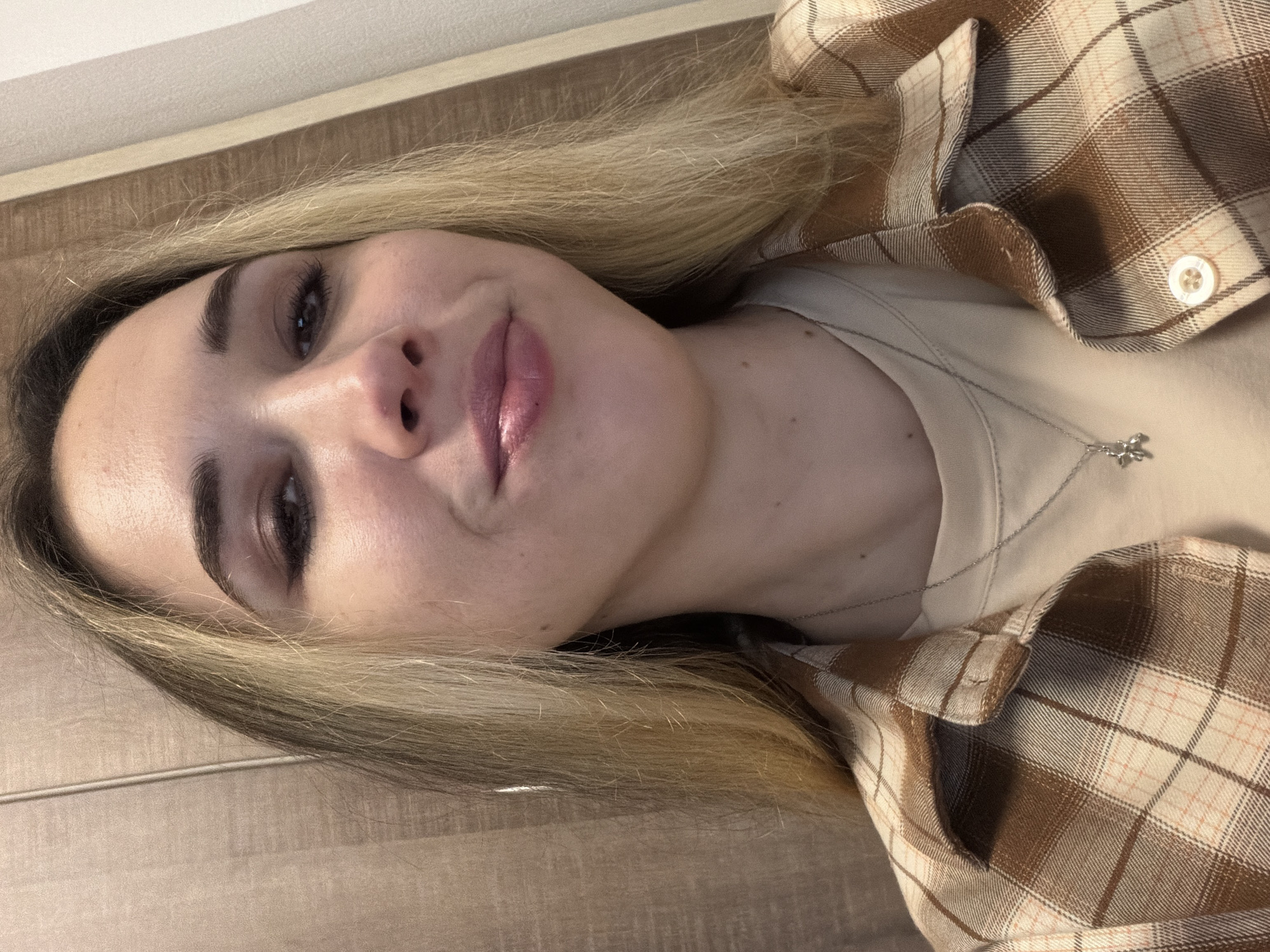 A close-up selfie of a young woman with fair skin, dark eyebrows, and dark eyes. She has dark hair at the roots that transitions into blonde highlights. She is wearing a light beige t-shirt and a brown and beige plaid shirt. A silver necklace with a small pendant is visible. The background is a wooden surface, possibly a wall or a floor. There is no information in the image to indicate its location as Rostov-on-Don, Russia.