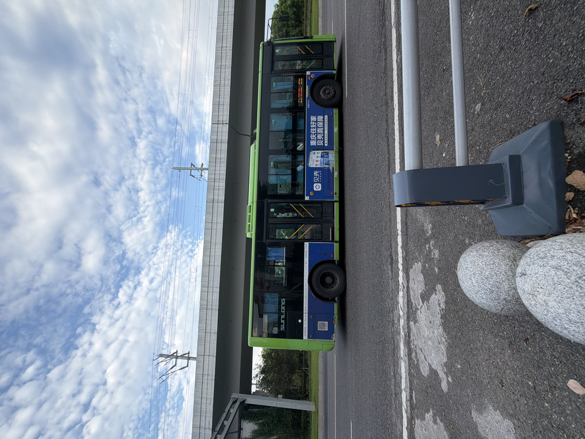 A green and blue electric bus, labeled "SUNLONG", is parked on an asphalt road under a concrete overpass with overhead power lines in Jiangjin, China. The bus has tinted windows and advertisements on its side, including one with Chinese characters and a smartphone icon. In the foreground, a gray metal guardrail is attached to a dark gray concrete base. Two large, speckled gray rocks are positioned behind the guardrail. The sky above is filled with scattered white clouds against a blue backdrop. The road has a white lane marking.