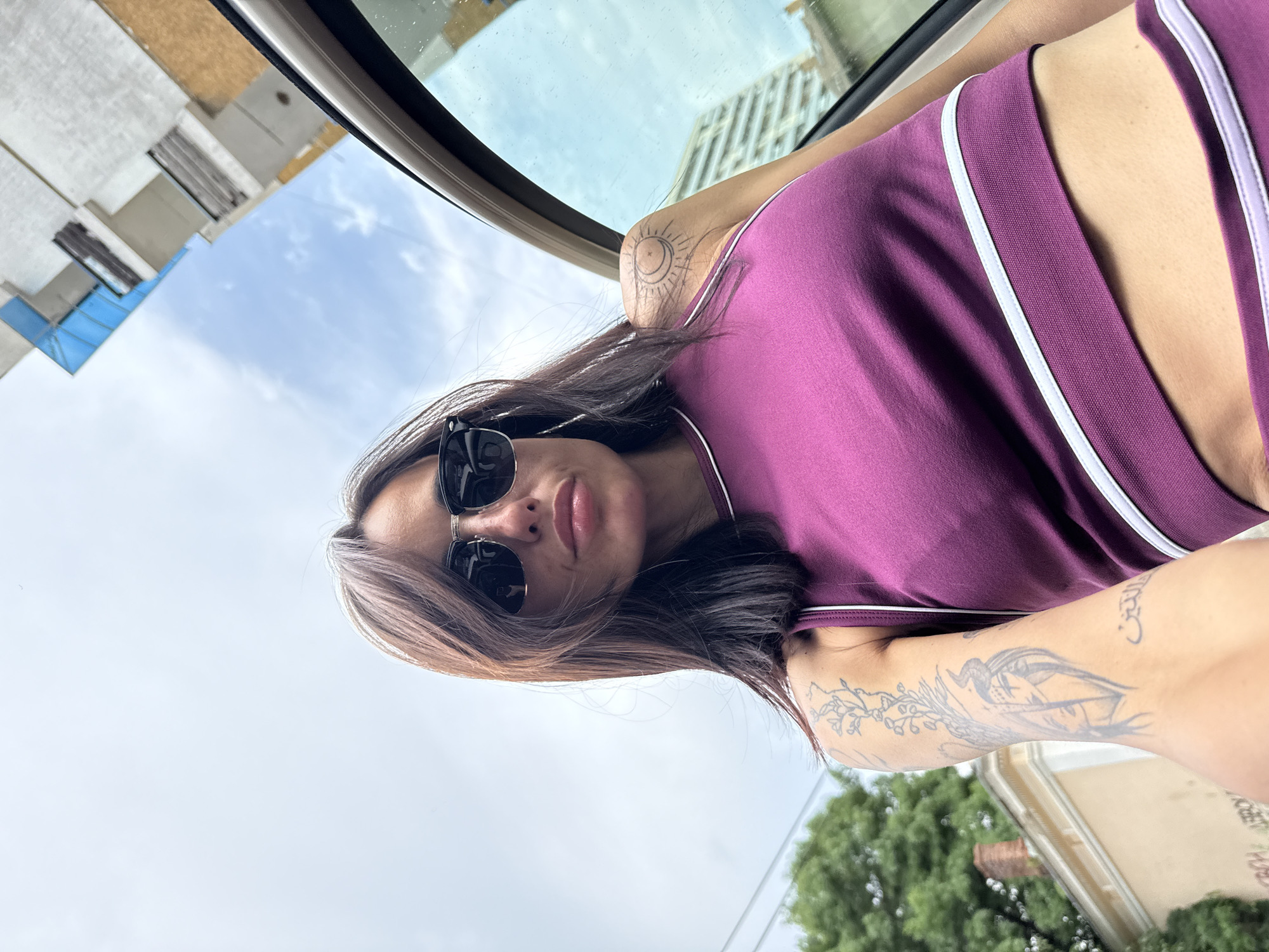 A medium-shot, low-angle, rotated selfie depicts a woman in sunglasses, wearing a purple tank top with white trim. She appears to be in a vehicle, likely a car, given the visible window frame and the backdrop of a cityscape. Her hair is brown with light streaks. A tattoo is visible on her left upper arm, and another on her right arm. The background shows buildings under a slightly overcast sky, suggesting a daytime scene. The location is Pleven, Bulgaria, as specified. Buildings, greenery, and sky indicate an urban environment with some vegetation.