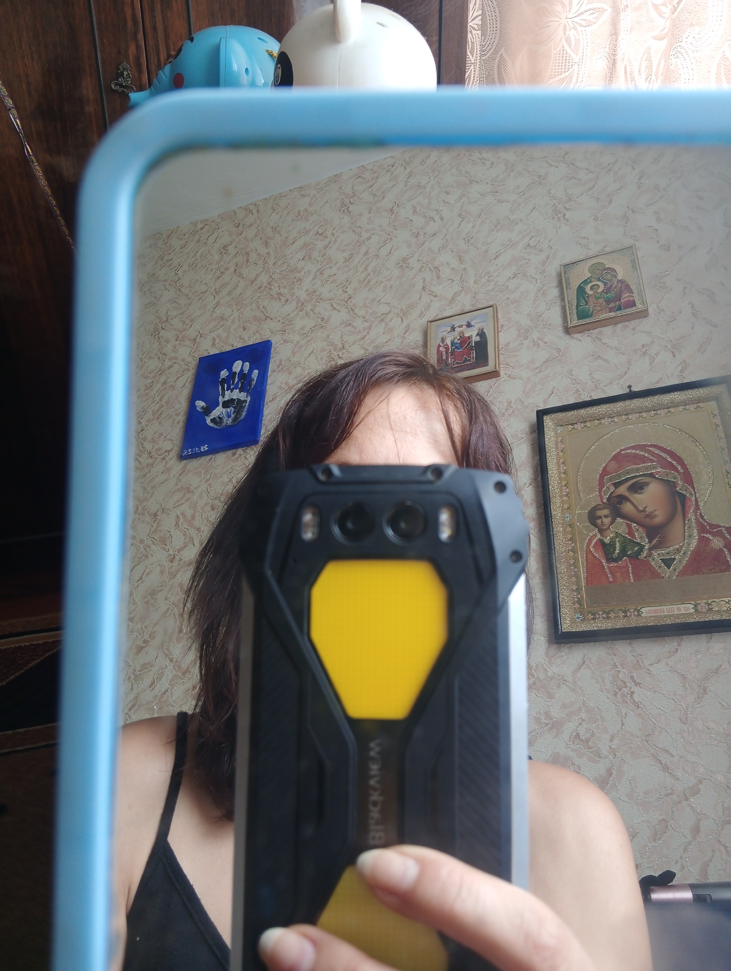 The image is a selfie taken in a room. A person is holding a black and yellow rugged smartphone in front of a mirror, reflecting their face and upper body. The background includes a textured wall with several religious icons and a blue-framed mirror. To the left, a wooden cabinet and a blue toy are visible. The room appears to be a private residence, likely a bedroom or living area. The city of Bar, Ukraine, is not discernible from the image itself; this information is external to the visual content.