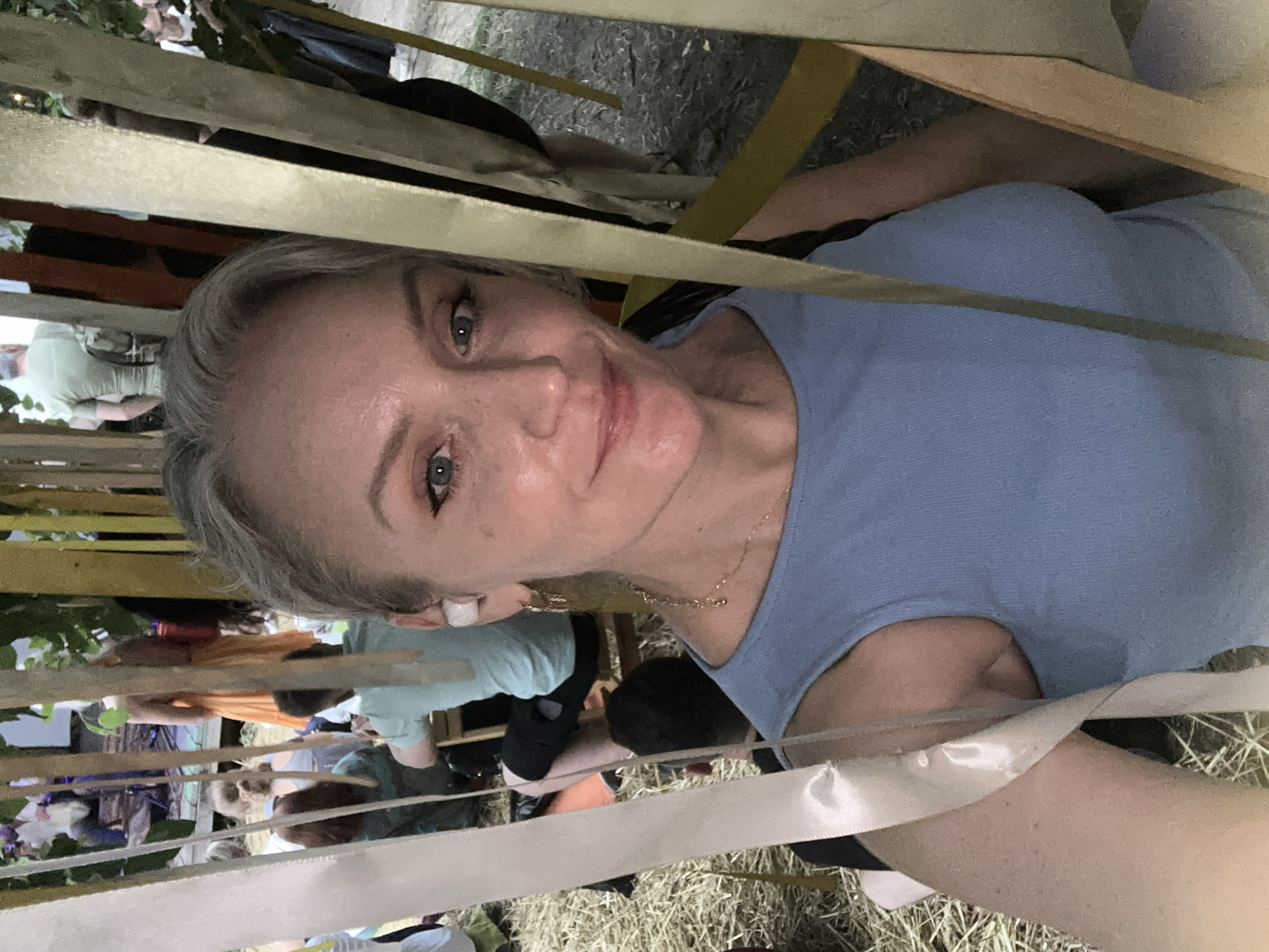 In this selfie, a woman with blonde hair, blue eyes, and light skin is positioned in the foreground, looking directly at the camera with a slight smile. She is wearing a light blue tank top and a gold necklace. A white earbud is visible in her left ear, and a gold dangle earring hangs from her left earlobe. The background is out of focus and appears to be an outdoor event. Multiple people are visible, some seated and some standing, in what looks like a gathering or festival. Some wooden structures and colorful fabric streamers are present, adding to the festive atmosphere. The ground is covered with hay or straw. The overall lighting suggests it is daytime. The context provided places this scene in the city of Kyiv, Ukraine.
