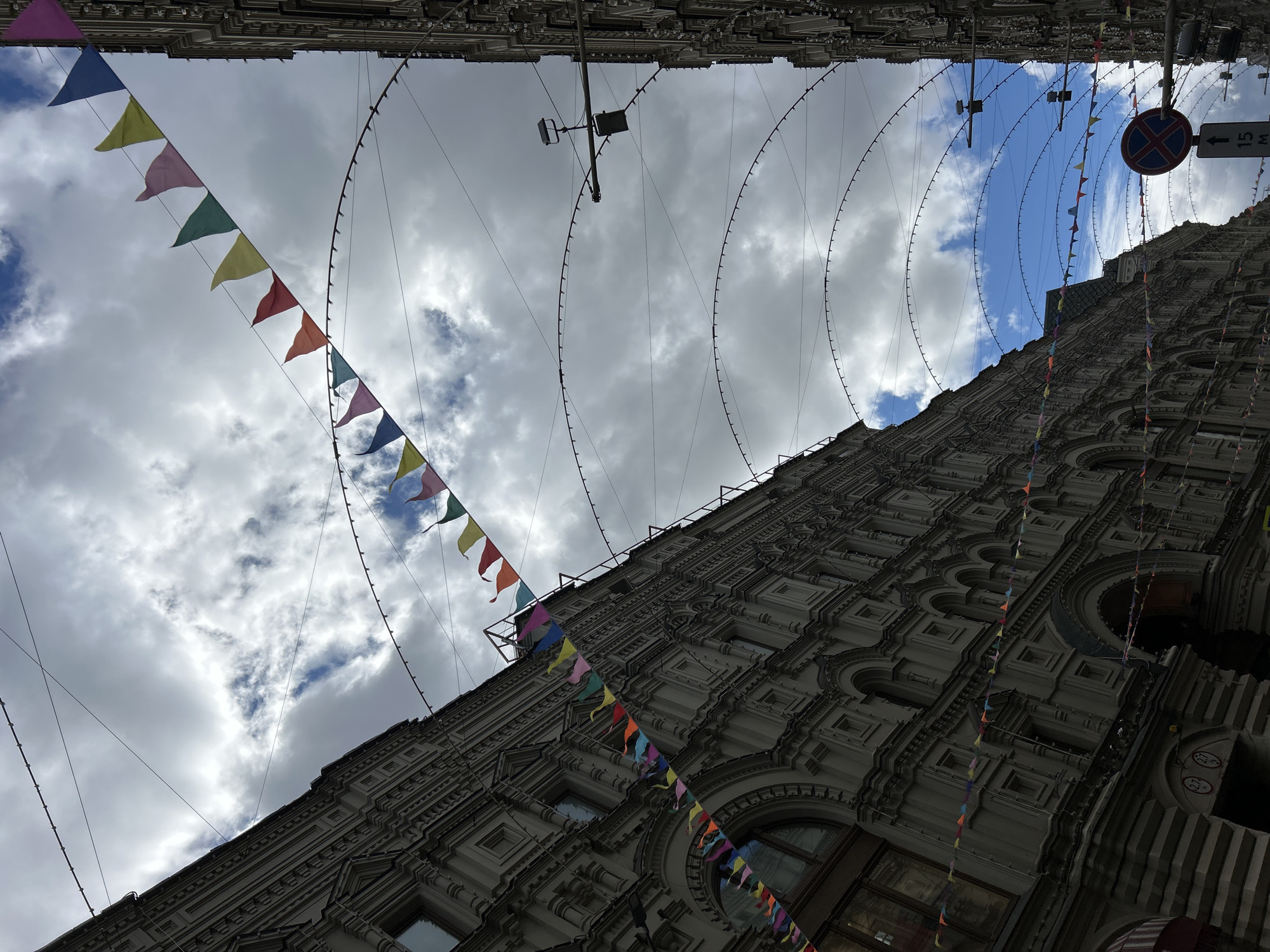 A low-angle shot captures a street scene in an unidentified city. The architecture features a detailed facade with windows and moldings, contrasted by a sky with variable cloud cover. Colored triangular pennants are strung overhead, adding festive elements. The scene includes a road sign, indicating street regulations. Street lighting and other infrastructure are apparent.