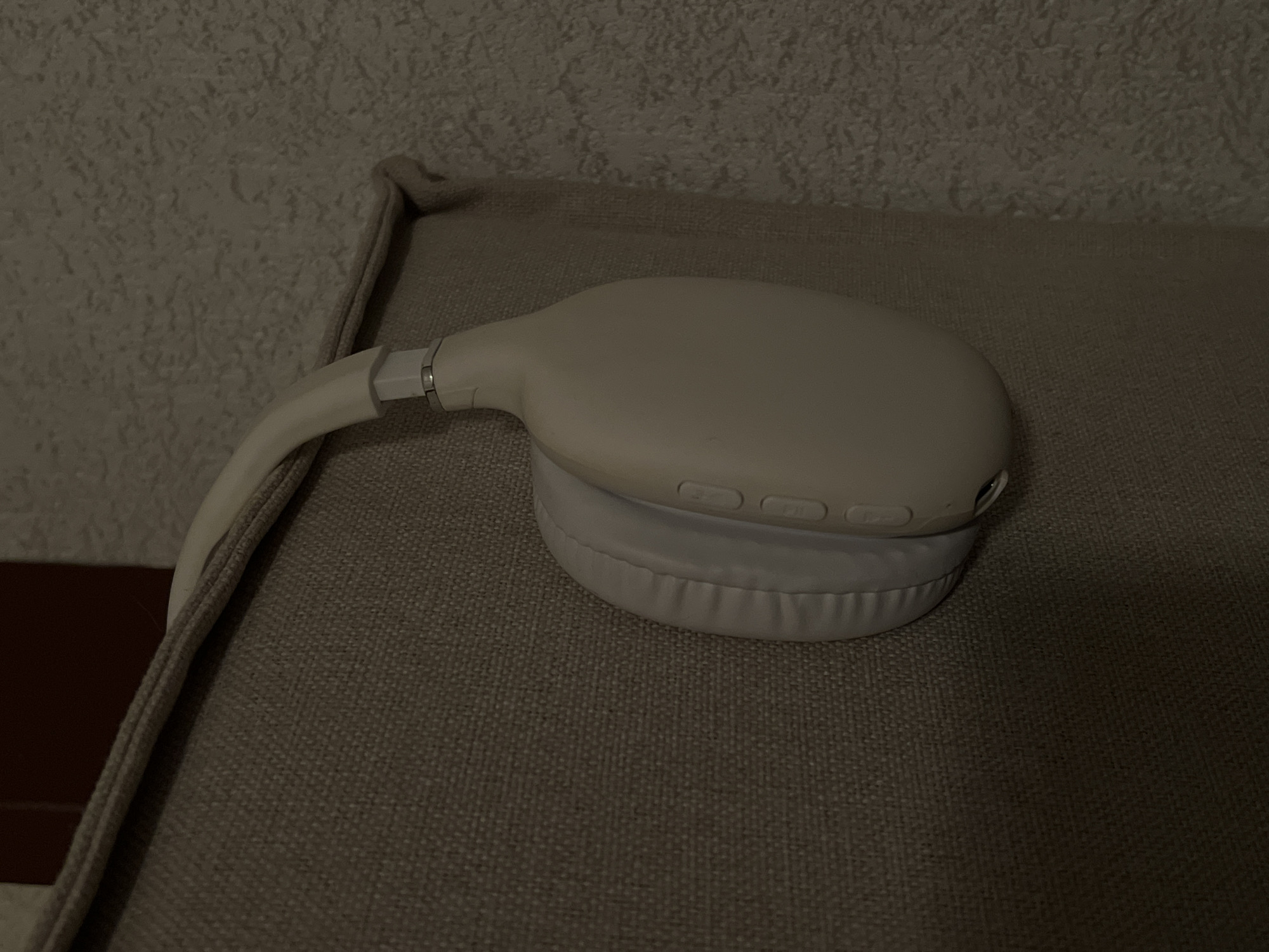 An off-white pair of over-ear headphones are resting on the edge of a light brown upholstered piece of furniture. The headphones are positioned so that one earcup is visible, with its padded surface and three buttons on the side. The headband arches over the top of the furniture. The background consists of a textured wall. The scene appears to be indoors, in a dimly lit room. The location in Genemuiden, Netherlands is not discernible from the image.