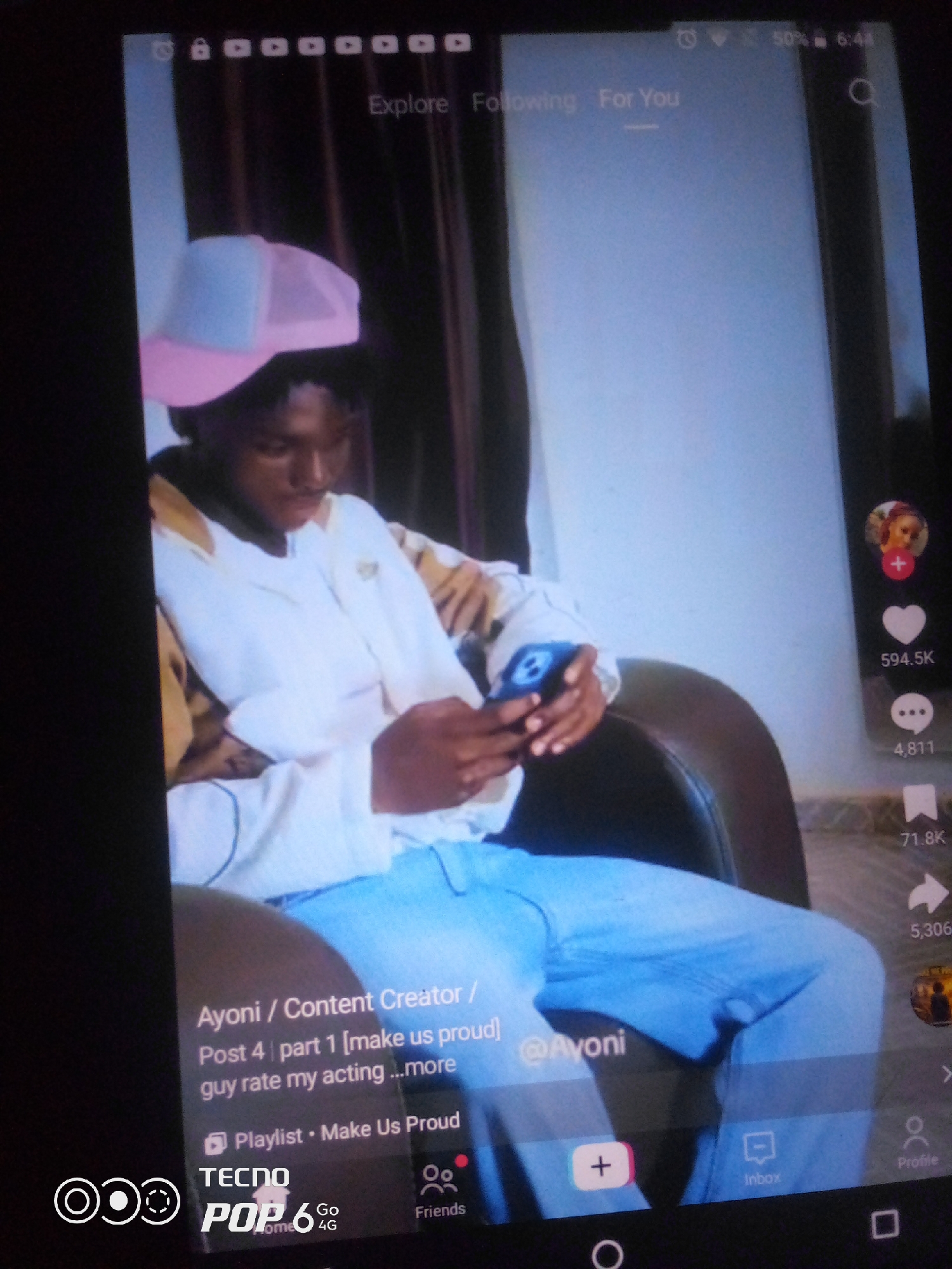 The image shows a close-up of a person sitting in a chair, looking at a phone. The person is wearing a white shirt and blue jeans, and a pink and blue baseball cap. The phone is displaying a social media app, possibly TikTok, with a video of the person playing. The user is holding the phone in their hands and appears to be watching the video. The background is blurred but appears to be an indoor setting with curtains. The bottom of the image shows the TECNO POP 6 logo. There are no discernible landmarks or contextual clues within the image to suggest a specific location in the city of Howayi, Nigeria.