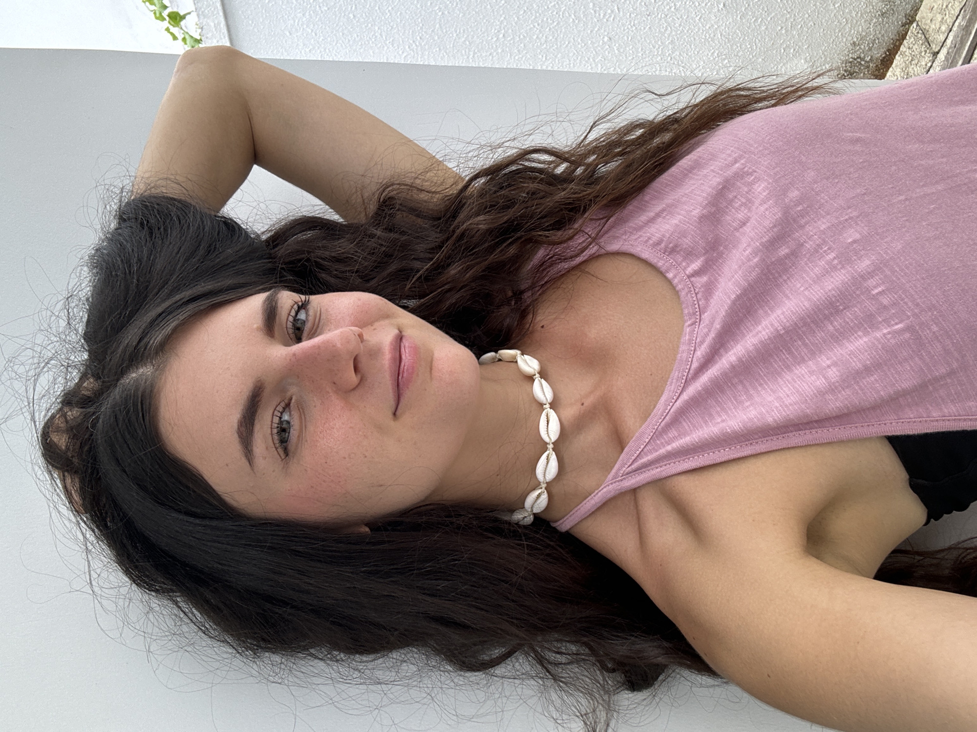 A young woman is depicted lying down, looking directly at the camera with a soft, relaxed smile. She has long, dark, wavy hair cascading around her head and shoulders, and fair skin with scattered freckles visible across her nose and cheeks. Her eyes are light in color, appearing grey or green.
She is dressed in a simple, light pink scoop-neck tank top, with a black bra strap or garment peeking out from under her left armpit. A prominent accessory is a necklace made of several small, white, cowrie-like shells worn around her neck. Her right arm is bent, with her hand positioned near her head, while her left arm is also bent, resting beside her body.
The setting appears to be primarily a plain, light-colored surface, possibly a white wall or floor. In the upper right corner, a textured white surface is visible, along with a hint of green foliage and a faint outline of what could be an outdoor railing or structure, suggesting an outdoor balcony or patio. Given the location context of Mũi Né, Vietnam, this fits a tropical or coastal environment.
The lighting is bright and even, typical of daytime conditions, casting no harsh shadows. The overall impression is one of calm and contentment. The image is presented in a landscape orientation, with the woman lying horizontally across the frame. There is no visible text.