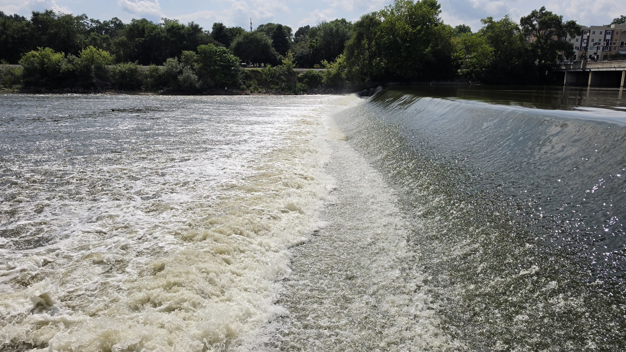 The image depicts a river with a dam in the foreground. The river water on the left side of the dam is turbulent and frothy, indicating a strong current. On the right side, the water flows smoothly over the dam, creating a waterfall effect. Lush green trees line the riverbank in the background, and a bridge with buildings can be seen in the distance. The sky is partly cloudy, with patches of blue visible. The scene appears to be a natural landscape with some man-made structures. The location is identified as the Fox River in Elgin, Illinois.