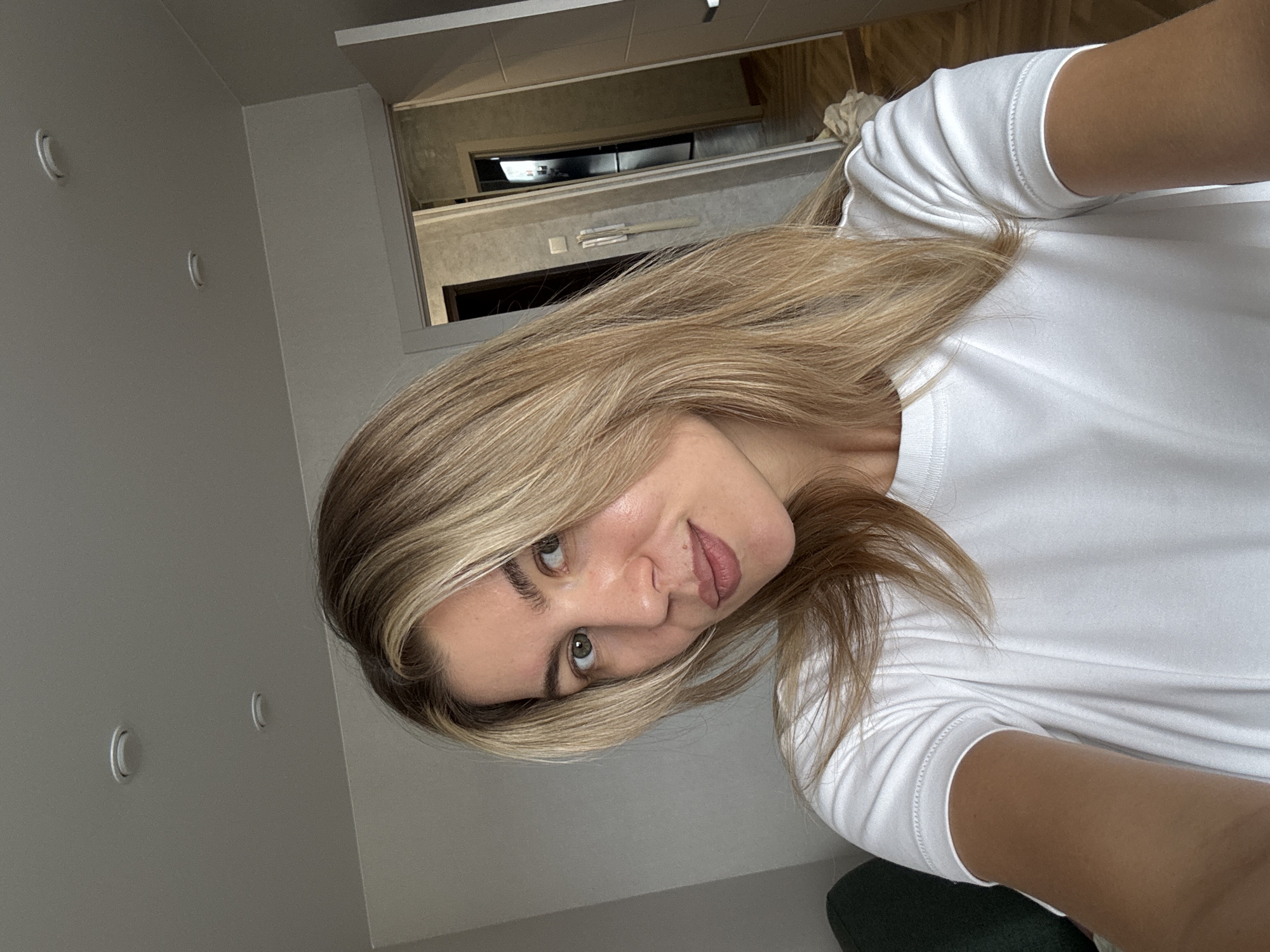A medium close-up, tilted selfie captures a young woman indoors. She has long, wavy blonde hair with lighter highlights, fair skin, and light-colored eyes, which are directed towards the viewer. Her lips are slightly parted in a subtle smile, conveying a gentle and approachable expression. She is wearing a simple, white short-sleeved t-shirt.
The background consists of plain, light-colored walls. In the upper right quadrant, an open doorway or window provides a glimpse into another interior space, possibly a hallway or adjacent room, featuring a grey ceiling and indistinct architectural details. On the left wall, two small, C-shaped light fixtures or hooks are visible. A dark, possibly green or blue, object (perhaps a piece of furniture) is partially visible in the bottom right corner, beneath her arm. The lighting is bright and even, illuminating her face and the immediate surroundings. No text is visible in the image.