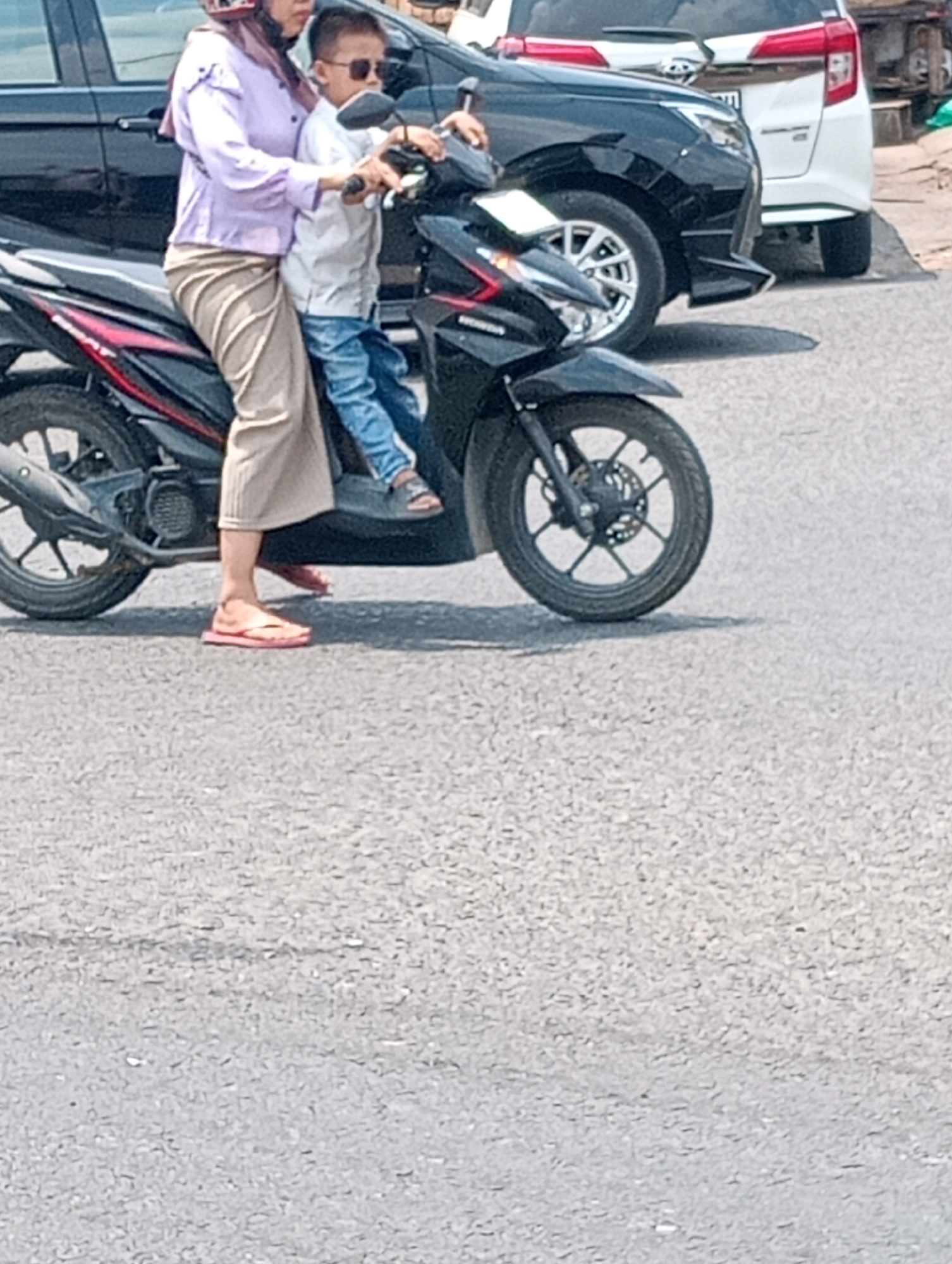 A woman wearing a light purple shirt, beige pants, and a hijab is riding a black motorcycle. She is wearing pink flip-flops. A young boy, wearing a white shirt, blue jeans, and sunglasses, is sitting in front of her on the motorcycle. They are on a paved road, likely in Bandar Lampung, Indonesia, with a black car and a white car visible in the background. The image appears to be taken during the daytime.