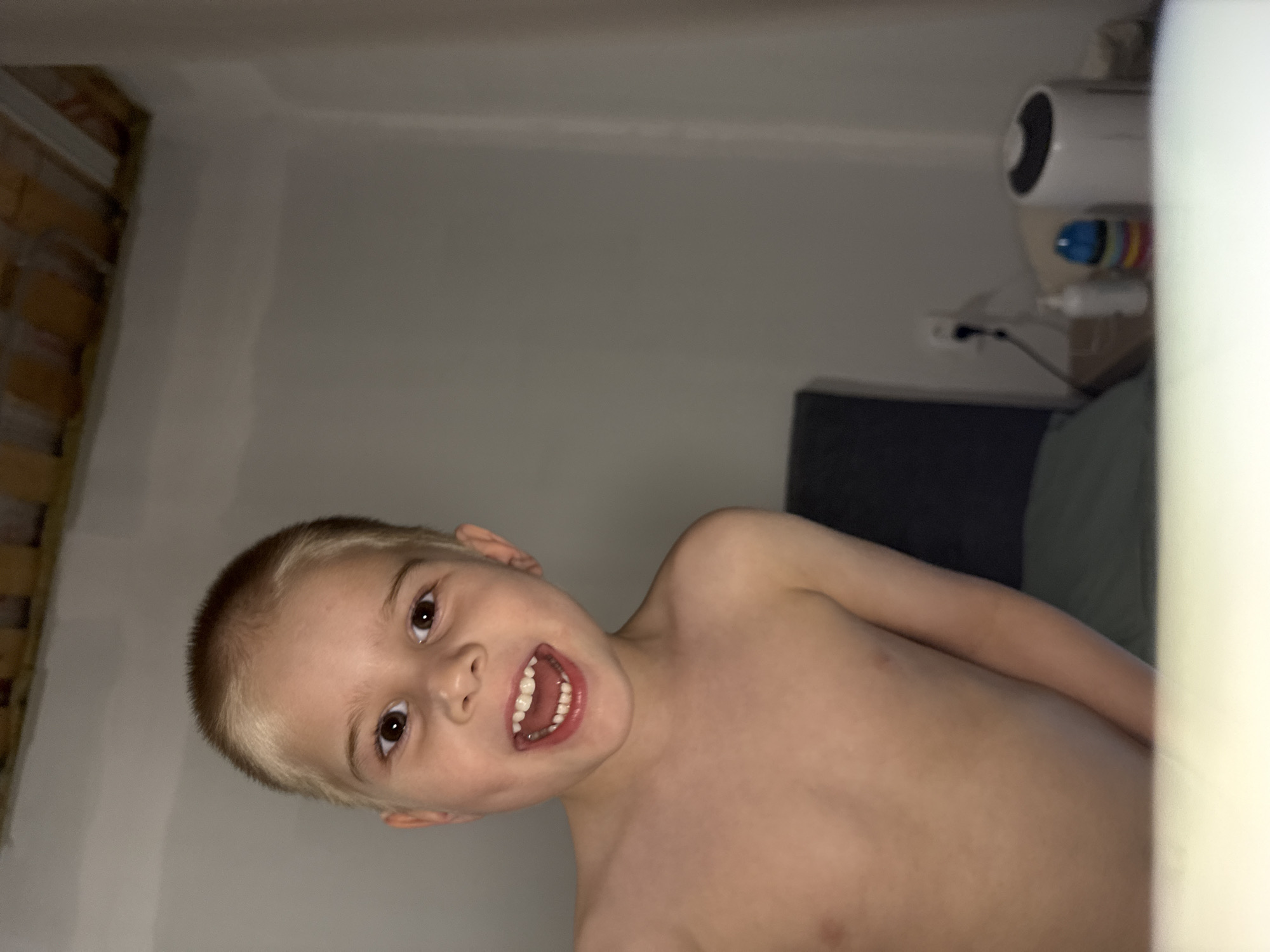 A blond, shirtless boy with brown eyes and visible teeth is posing for a photo indoors. The background is a plain, light-colored wall. Visible behind him is a dark gray headboard and what appears to be a humidifier with other small objects on a shelf. The image contains no verifiable data regarding the city of Chaikovsky, Russia.