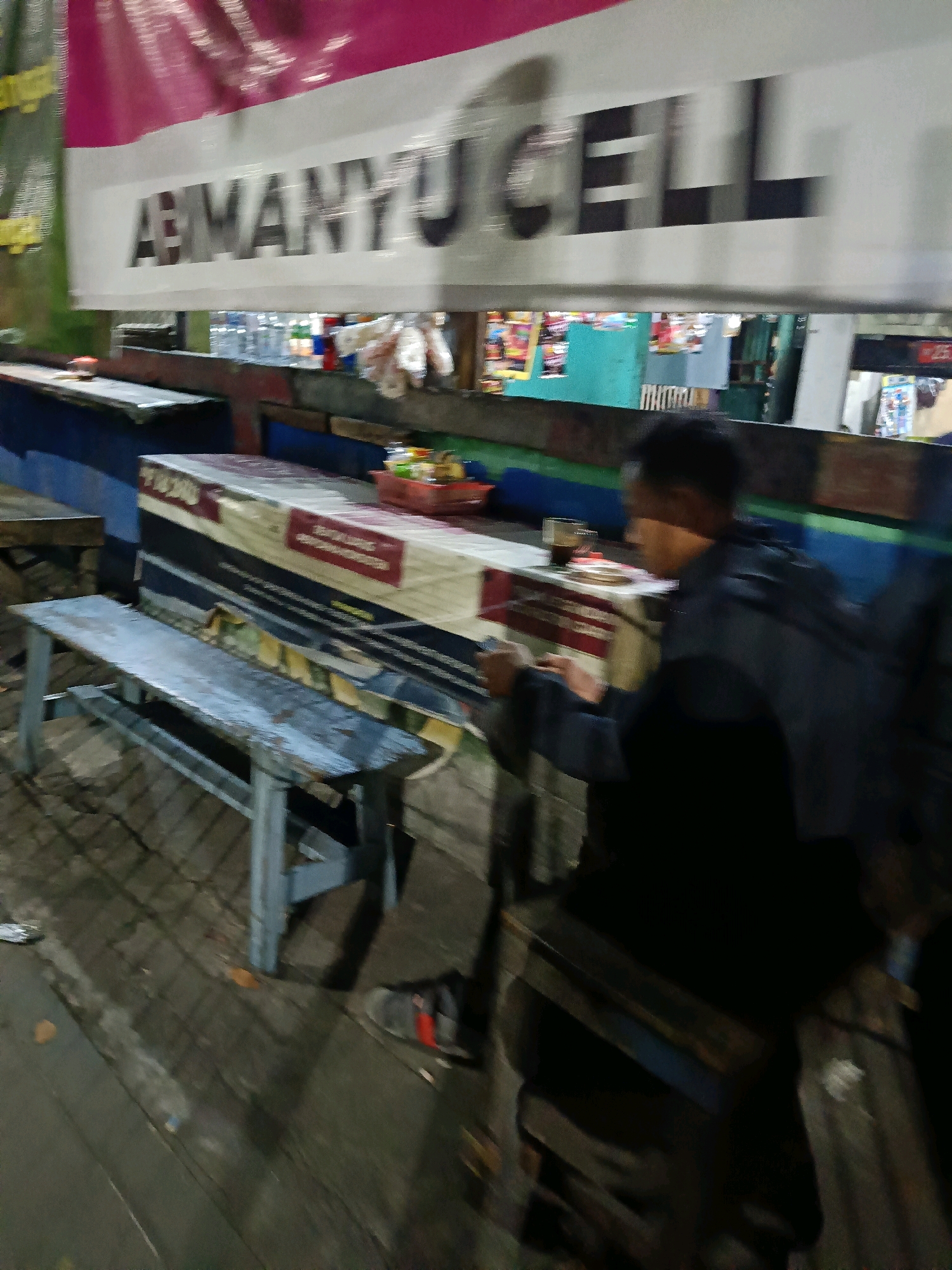 In Kedungrejo, Indonesia, a man sits at a table in what appears to be an outdoor seating area, possibly a roadside stall or a small shop. He is looking down at his phone, his face obscured by shadow and the blur of motion. The man is wearing a dark jacket over a black shirt.
In the background, a banner with the text "ABIMANYU CELL" is visible, suggesting the presence of a mobile phone service provider. There are also shelves stocked with various items behind the banner, indicating a retail space. The overall environment suggests a commercial area, perhaps a market or a street with shops.
The image is taken at night, with artificial lighting creating a somewhat blurry and atmospheric scene. The wooden floorboards and simple benches contribute to a casual, unpretentious setting.