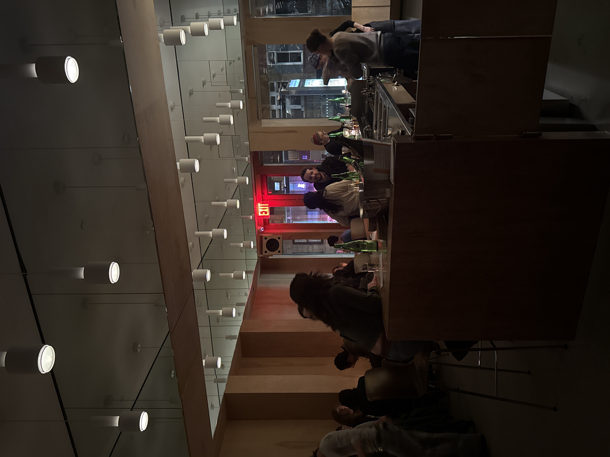 The image depicts the interior of a bar or restaurant, likely in New York City, based on the urban architecture visible through the windows. Patrons are seated at a long bar counter. A bartender is visible behind the counter, engaging with customers. Several people are sitting at the bar counter, appearing to socialize. Empty beer bottles are seen. The lighting in the bar is a mix of warm, ambient light and cooler, brighter spotlights. The ceiling features a patterned arrangement of cylindrical light fixtures. The color red appears to be a design element within the space, noticeable on the exit sign.