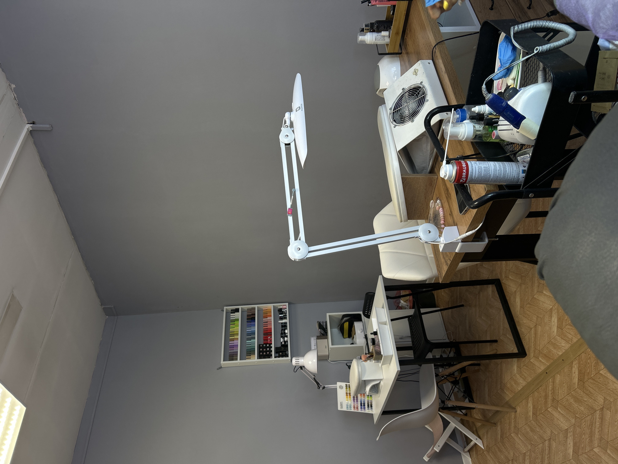This image depicts an organized indoor workspace, likely a nail salon or dedicated nail art studio, viewed from an elevated perspective and rotated ninety degrees. No people are visible in the scene.
The primary focus is on two distinct workstations. The one closer to the viewer features a large, white, adjustable desk lamp with a flat, rectangular head, positioned over a wooden tabletop. To its right, a white ergonomic chair with a high back is pulled up to the table. On the table itself, there's a white square nail dust collector with a metal grill. Adjacent to it, a black metal rolling cart holds various bottles, including spray cans (one prominently displays "BRAUBERG" and "SUPER FIX" in red text, another partially shows "CONSTANT"), liquid dispensers, and a coiled wire. Blue gloves are also visible within the cart.
Further into the room, a second, smaller workstation consists of a white table with a compact desk lamp. Above this table, a white wall-mounted shelf unit displays numerous small bottles, presumably nail polishes or gels, arranged neatly by color. On the table itself, there's an organizer filled with colorful nail polish swatch sticks. A light-colored chair, possibly white plastic, sits next to this table.
The walls of the room are painted a solid medium grey. The floor features a wooden parquet pattern, possibly a herringbone or chevron design, creating a textured surface. The space appears well-lit by the desk lamps, suggesting a functional and tidy environment for performing nail services. The time of day cannot be determined, but the artificial lighting suggests it could be any time.