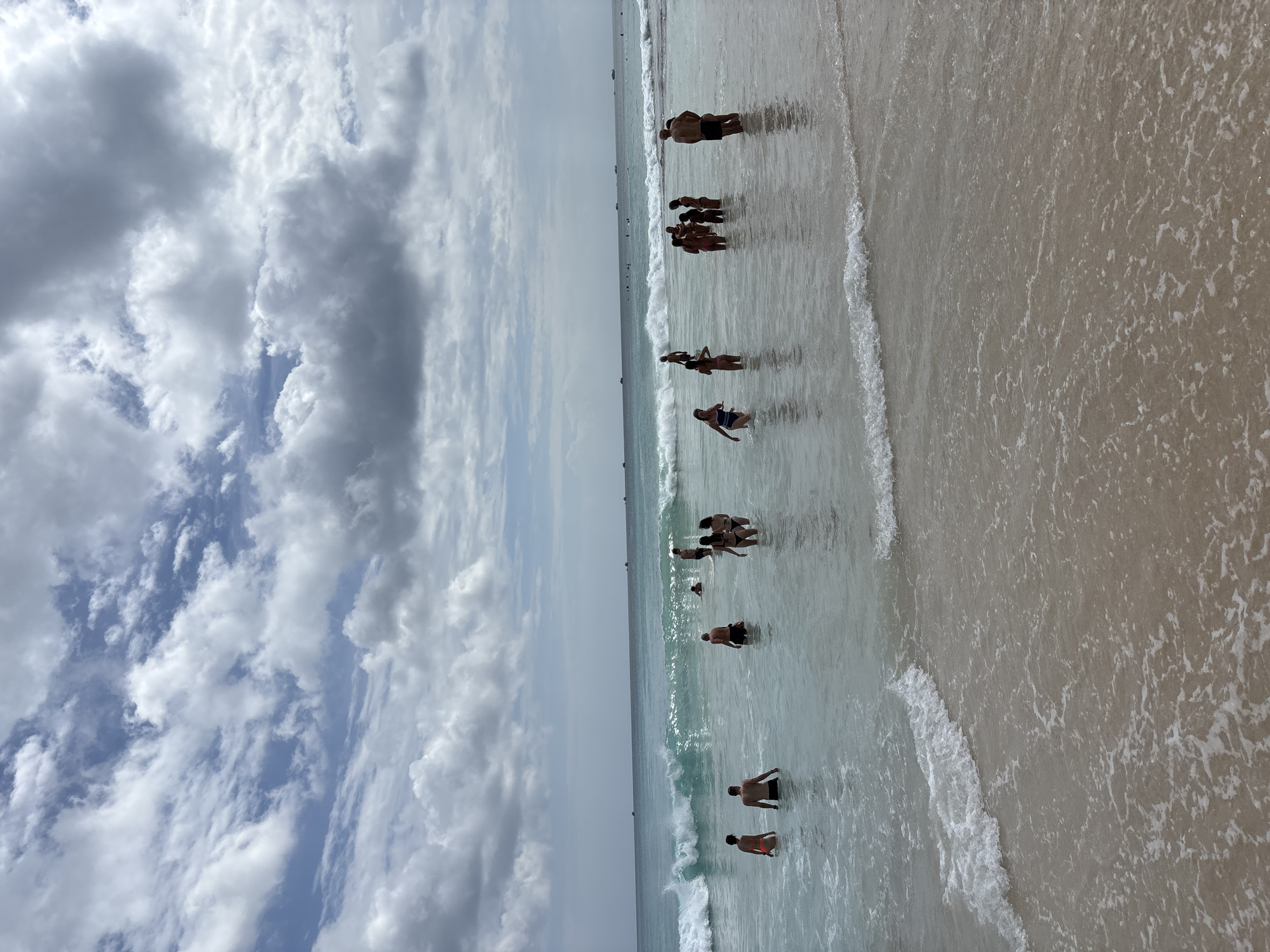A group of people are wading in the shallow waters of a beach in Kuta Selatan, Indonesia. The water is clear and calm, with small waves breaking on the shore. The sky is cloudy, with patches of blue visible between the clouds. In the distance, several boats are visible on the horizon. The people are dressed in swimwear and appear to be enjoying themselves. Some are standing in the water, while others are walking along the shore. The overall impression is one of a relaxed and pleasant day at the beach.
