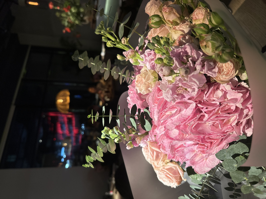 The image displays a vibrant bouquet of flowers, oriented vertically as if viewed from above and slightly to the side. The bouquet is the clear focal point, filling a significant portion of the frame, with a shallow depth of field rendering the background softly blurred.
The dominant flowers in the bouquet are large, densely petaled blossoms in various shades of pink, ranging from a rich, bright pink to lighter, more delicate pastels. These appear to be hydrangeas or a type of full-blooming rose like garden roses, characterized by their numerous ruffled petals. Interspersed among these are smaller, pale pink or peach-colored roses and rosebuds, some still tightly closed. Clusters of delicate, light pink or lavender flowers, possibly stock or similar tall-stemmed blooms, add texture and height.
Complementing the blossoms is ample green foliage, most notably distinctive eucalyptus leaves with their rounded, slightly silvery-green appearance, adding a natural and elegant touch. There are also slender green stems with developing buds, contributing to the bouquet's fresh look. The flowers are arranged compactly, suggesting they are wrapped, with a portion of light-colored, possibly white or cream, wrapping material visible along the right and bottom edges of the bouquet.
The background is dark and heavily out of focus, indicating that the photo was taken indoors at night or in a dimly lit environment, possibly near a window. Blurred lights create soft orbs of color: a prominent, elongated yellow or orange light is visible in the middle-left, alongside streaks of red and blue lights further down. These could be reflections, neon signs, or distant city lights viewed through glass. A faint outline of what appears to be a window frame or structural element can be discerned in the dark background. Another blurry hint of a floral arrangement or plant is visible in the far upper-left corner of the background, also out of focus.
No discernible text is visible in the image. The overall impression is one of beauty and elegance, focusing on the fresh and colorful floral arrangement against a serene, dark backdrop.