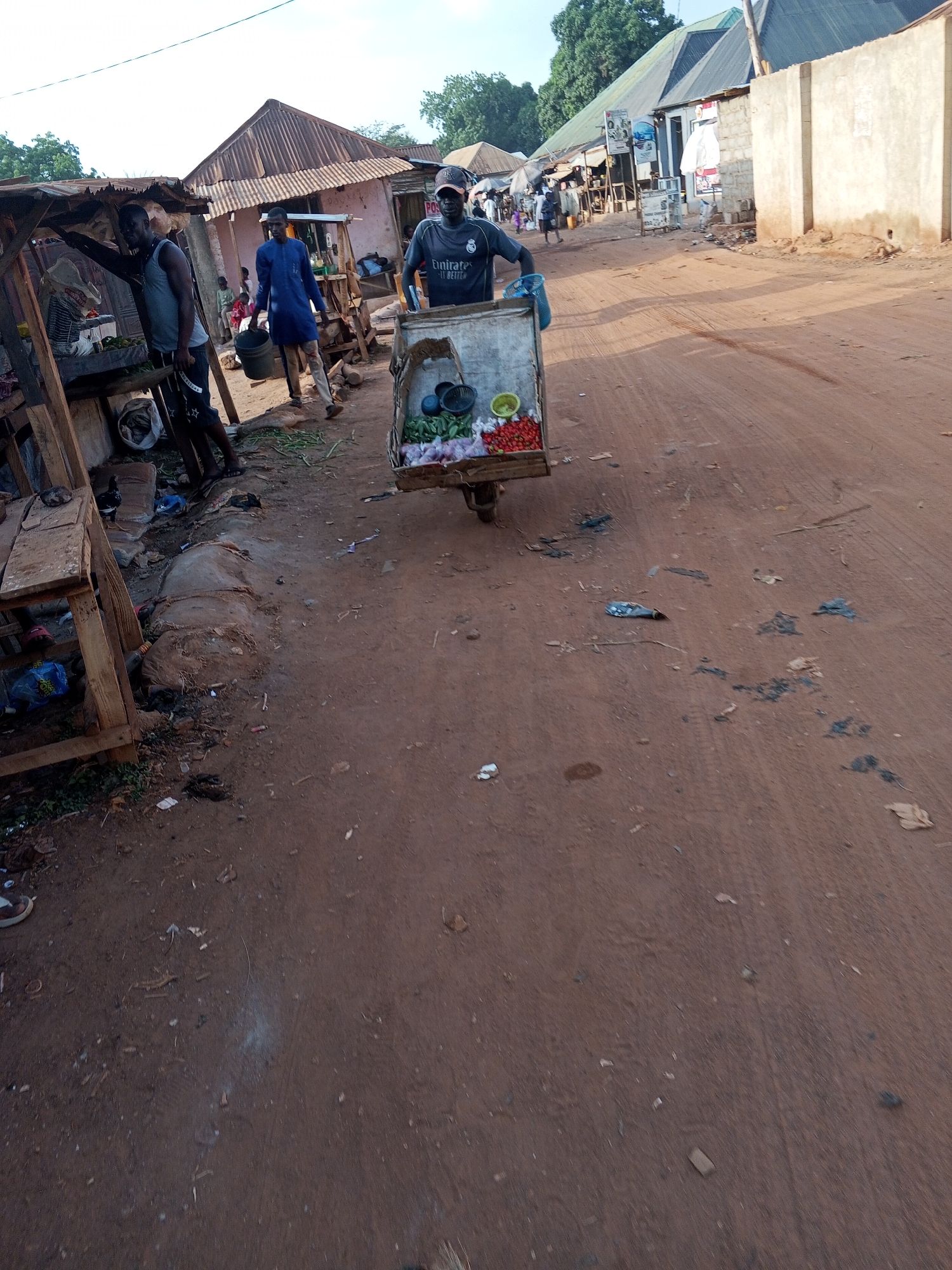 The image depicts a street scene in a market area of Abuja, Nigeria. The ground is predominantly red dirt. A man wearing a face mask and a gray Emirates t-shirt pushes a cart containing produce, including green vegetables, tomatoes, and possibly other items, down the street. Several other individuals are visible, including men standing near a wooden structure or stall on the left side of the frame. One man is carrying a bucket. Buildings with corrugated iron roofs are visible in the background. Trash and debris are scattered on the ground. A weathered wall is present on the right side of the image. The environment appears to be a bustling, informal market area.