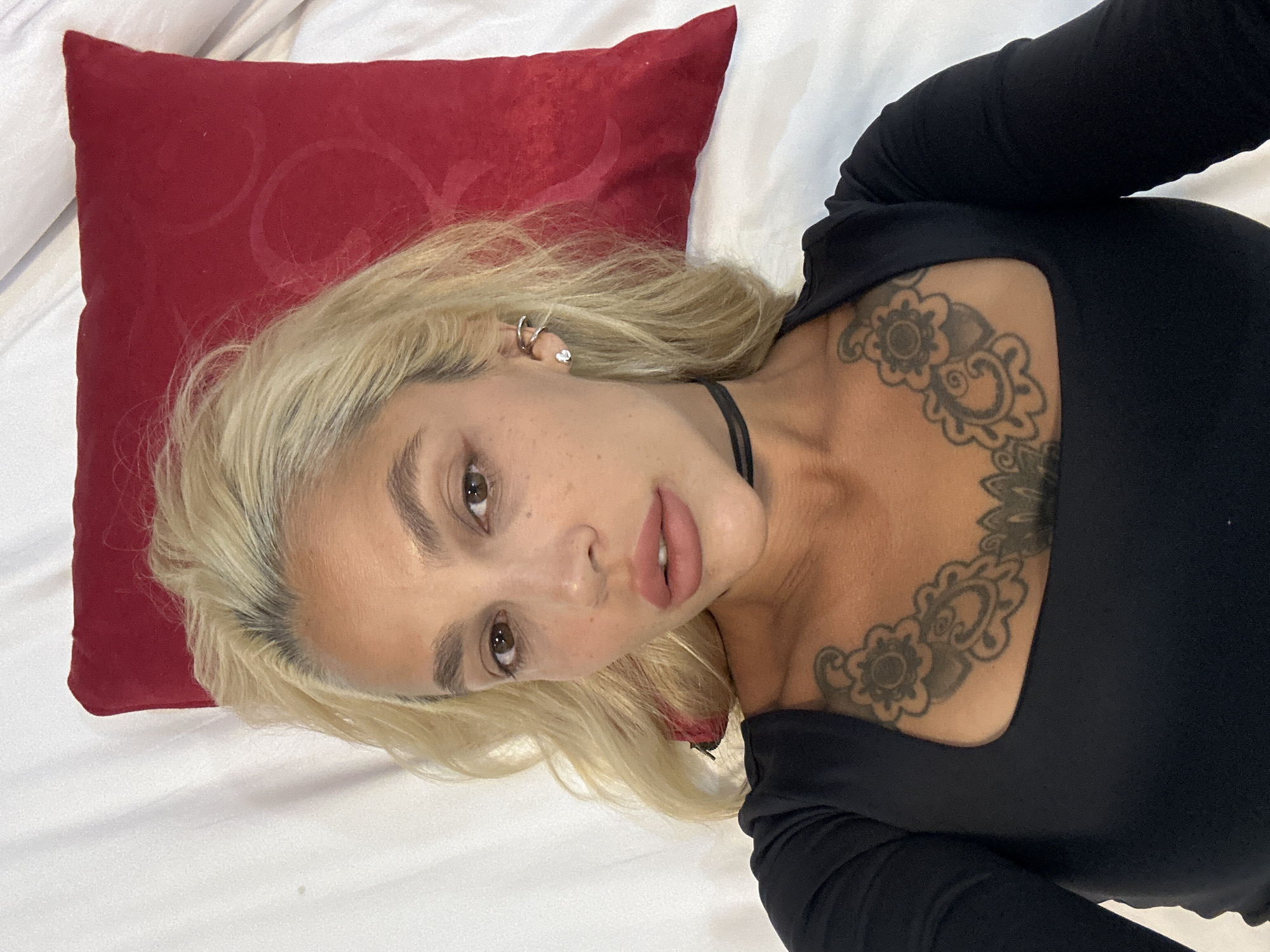 A woman with blonde hair and tattoos on her chest lies on a white surface, with a red pillow behind her head. She is wearing a black long-sleeved shirt and a black choker. The image appears to be taken indoors, possibly in a bedroom. There is no indication of the location being in Kuta Selatan, Indonesia.