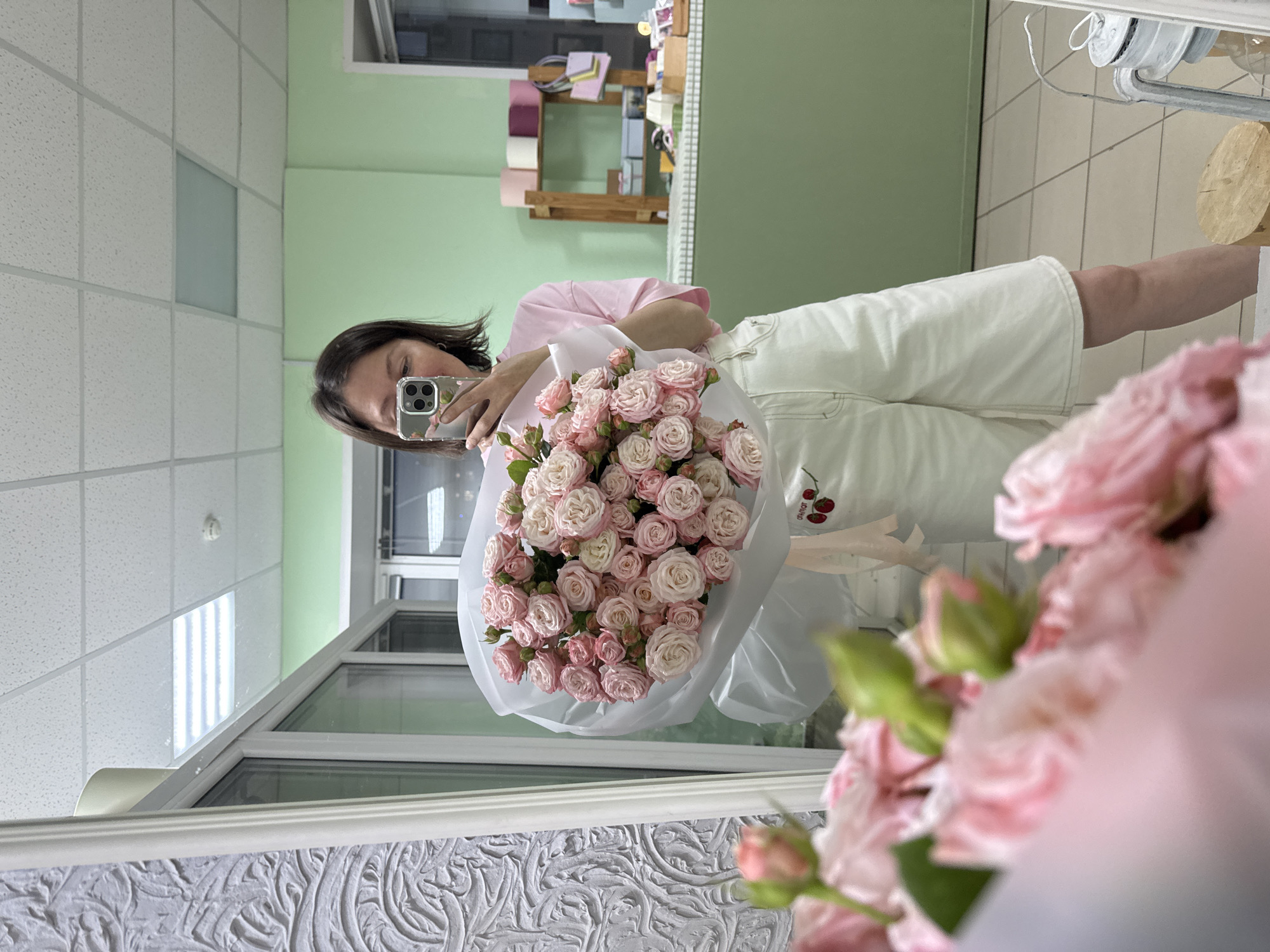 A young woman with brown hair is taking a selfie in a mirror while holding a large bouquet of pink roses. She is wearing a pink t-shirt and white shorts. The bouquet is wrapped in white paper and has a pink ribbon tied around it, with a small embroidered detail of cherries. The background appears to be a flower shop with shelves stocked with various items, including rolls of colored paper and small boxes. The walls are painted a light green, and there is a textured gray wall on the left side of the frame. The floor is tiled. The lighting suggests an indoor environment, possibly during the daytime. The overall scene is bright and cheerful.