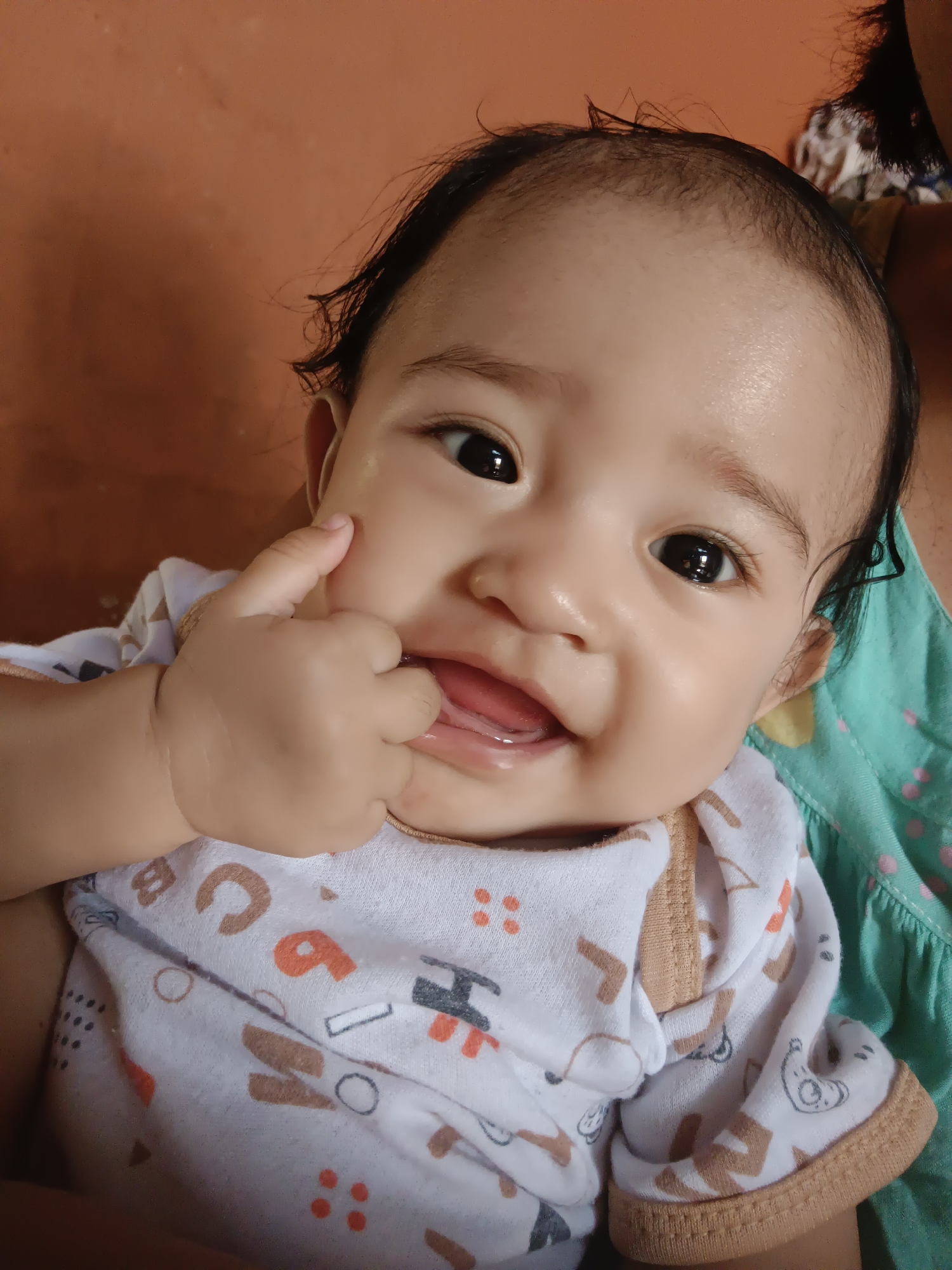 A close-up, eye-level shot shows a baby's face. The baby is looking directly at the camera with large, dark eyes and a slight smile. The baby's left hand is raised to its mouth, with the index finger touching the cheek and the thumb near the mouth. The baby is wearing a white onesie with an orange and brown alphabet print and tan trim. A portion of a green garment with pink polka dots is visible behind the baby. The background is a warm, orange-brown color. The scene appears to be indoors.