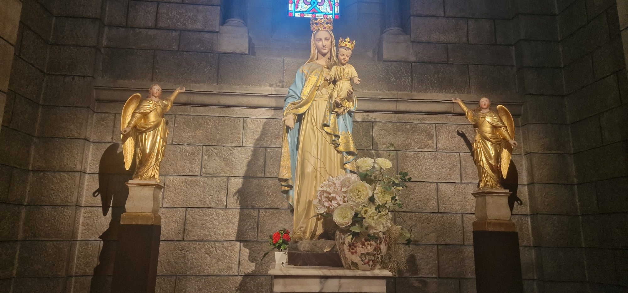 This is a full frontal, eye-level shot of a religious altar featuring a statue of the Virgin Mary holding the Christ child, flanked by two golden angel statues. The main statue depicts the Virgin Mary wearing a blue and gold robe and a crown. She is holding the infant Jesus, who also wears a crown. To the left and right of the central statue are two golden statues of angels, each with outstretched arms. In front of the Virgin Mary statue is a vase of light-colored flowers and a small pot of red flowers. The background is a stone wall. Above the statues, a stained-glass window is visible. This altar is located in the Saint Nicholas Cathedral in Monaco City, Monaco.