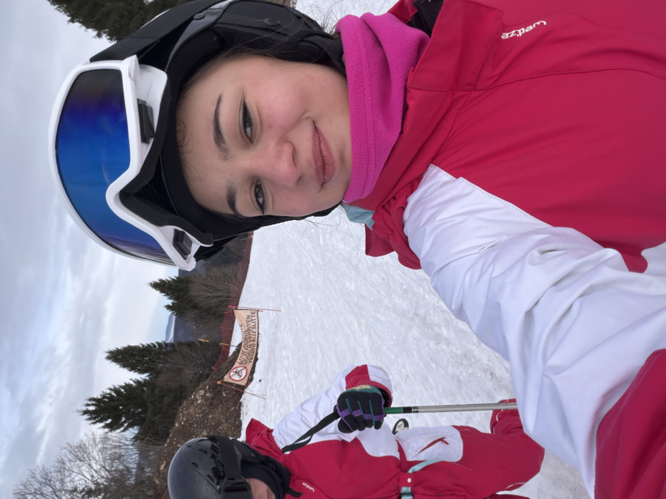 This image depicts a snowy ski slope scene, presumably located within the city of Belmont, France. No specific landmarks identifying Belmont are visible within the frame.
Two individuals are present on the slope. In the foreground, on the right, a young female is seen from the chest up, smiling towards the camera. She wears a black ski helmet with white and blue reflective goggles pushed up onto the helmet, a bright pink fleece neck gaiter, and a fuchsia and white ski jacket with white sleeves.
To her left, partially visible from the mid-torso down, is another individual. This person wears a black ski helmet, a dark neck covering, and a red and white ski suit. Their gloved left hand, with purple, green, and black accents, holds a ski pole.
The background features a snow-covered slope bordered by a brown, earthen bank on the left, behind which a red safety net is visible. A multi-lingual orange banner-like sign in the mid-ground displays a red circle with a crossed-out sled icon, indicating "No Sledding," with additional text including "VERBOTEN" (German) and "VIETATO ALLE SLITTE" (Italian). Coniferous trees populate the slopes in the background, with distant mountains visible beneath an overcast, grey sky.