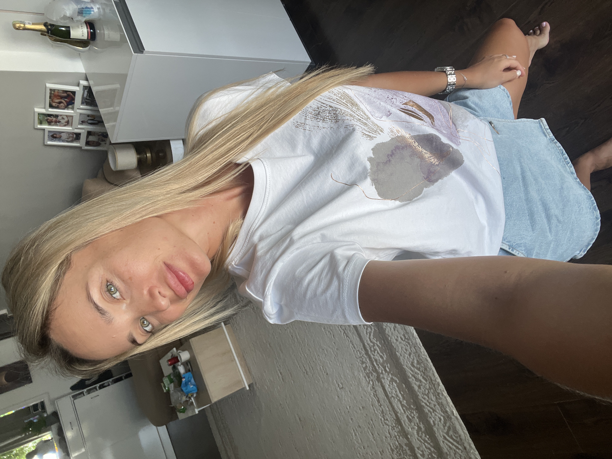 A blonde woman with green eyes is kneeling on a wooden floor in what appears to be a modern apartment. She is wearing a white t-shirt with a floral design and a light blue denim skirt. Her right hand is resting on her knee, and she is holding a phone in her left hand, taking a selfie. The background shows a living room area with a light-colored rug, a coffee table with items on it, and a white cabinet displaying several framed photographs and a bottle of champagne. The overall impression is of a casual, domestic setting. This scene is not specifically identifiable as being in Sharm El Sheikh, Egypt, but rather a generic interior space.