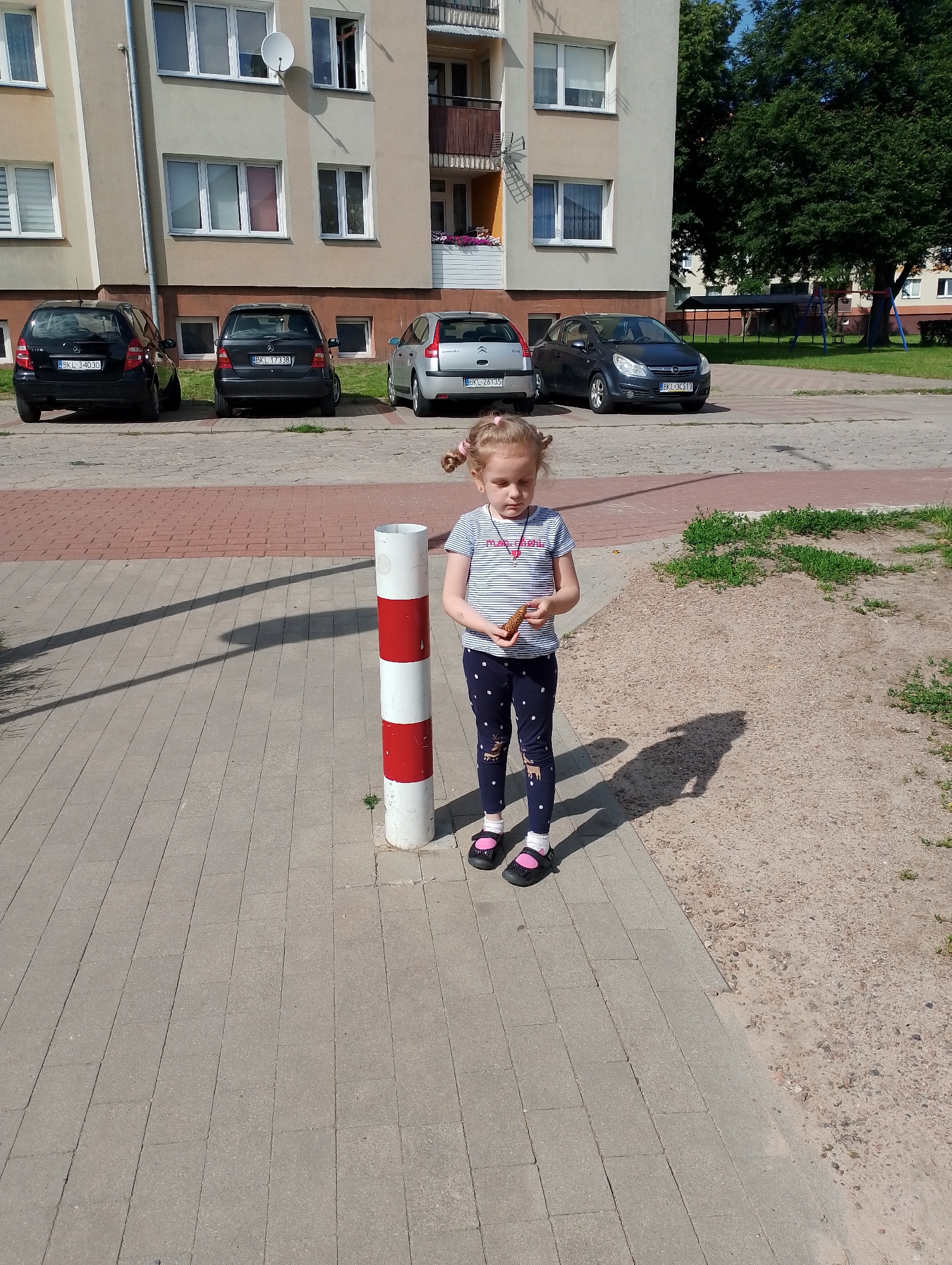 A young girl with blonde hair in pigtails stands on a paved walkway in front of a building. She is wearing a striped t-shirt, dark leggings with a white polka dot pattern and reindeer print, pink socks, and black shoes with pink accents. She holds a small snack in her hands and appears to be looking down at it. To her left, a red and white striped bollard stands on the walkway. In the background, several cars are parked in front of an apartment building. The ground to the right of the walkway is covered in gravel and sparse green vegetation. The sky is blue and the scene is illuminated by sunlight, casting shadows on the ground.