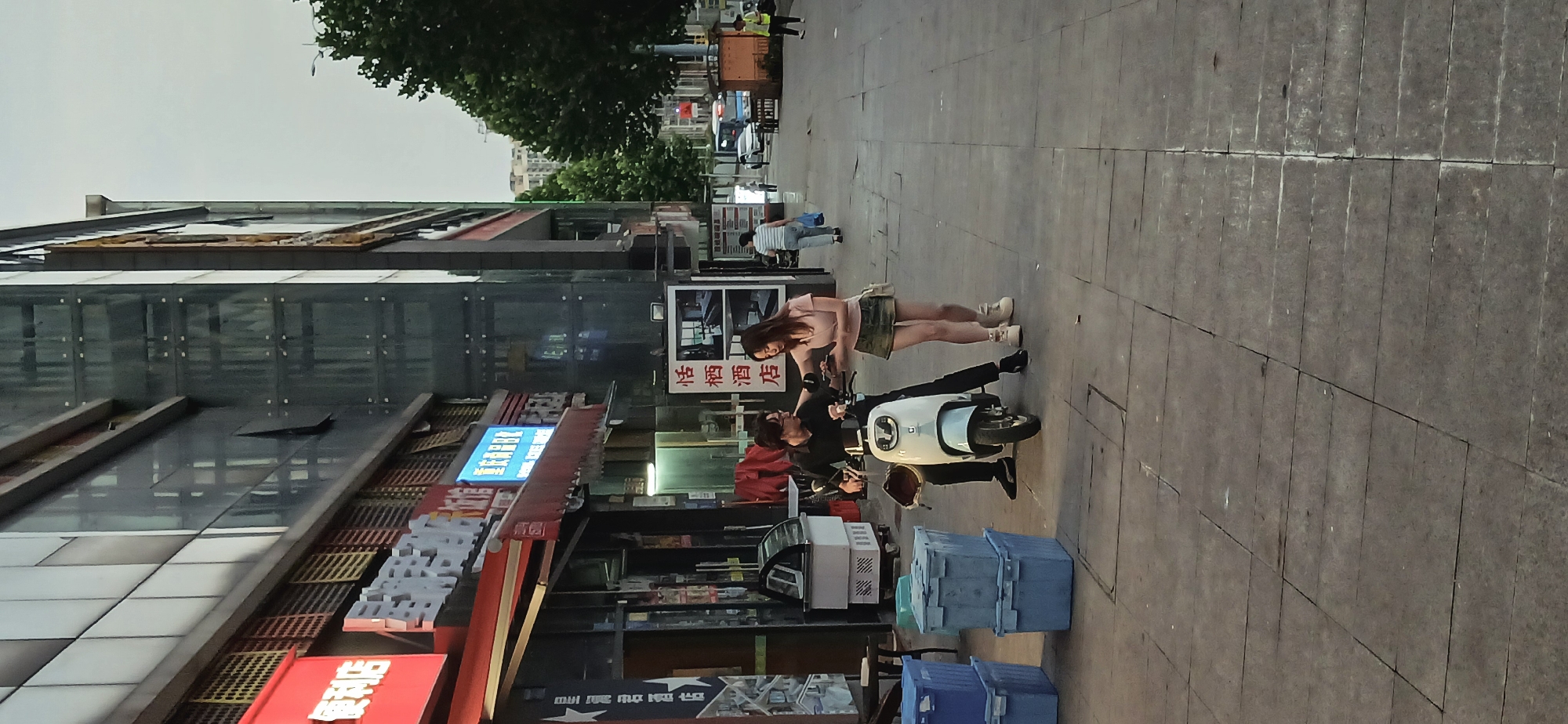 In Jiulongpo District, China, a man and a woman are positioned on a sidewalk next to a parked white scooter. The man is sitting on the scooter while the woman stands next to him. The sidewalk is paved with rectangular stone slabs. Buildings with commercial signage are visible on the left side of the frame. Other pedestrians are walking in the background.