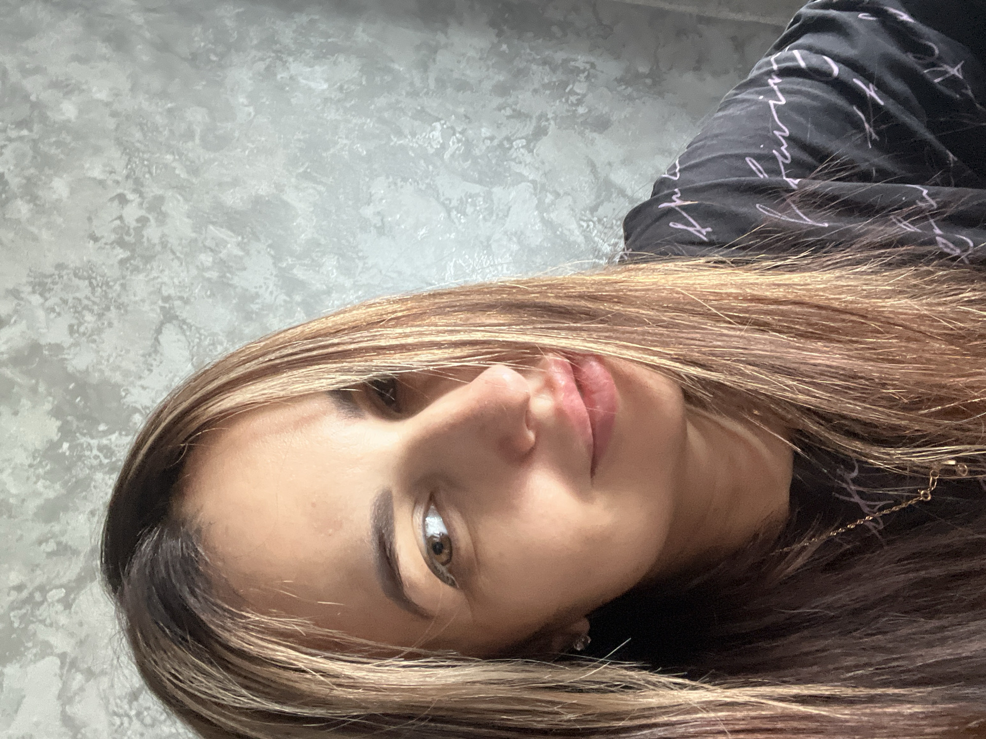 A woman is seen from a low angle, her face occupying much of the frame. She has fair skin, light brown hair with highlights, and brown eyes. She is wearing a black shirt with white writing and a gold chain. The background is a gray wall. The city and location are unknown.