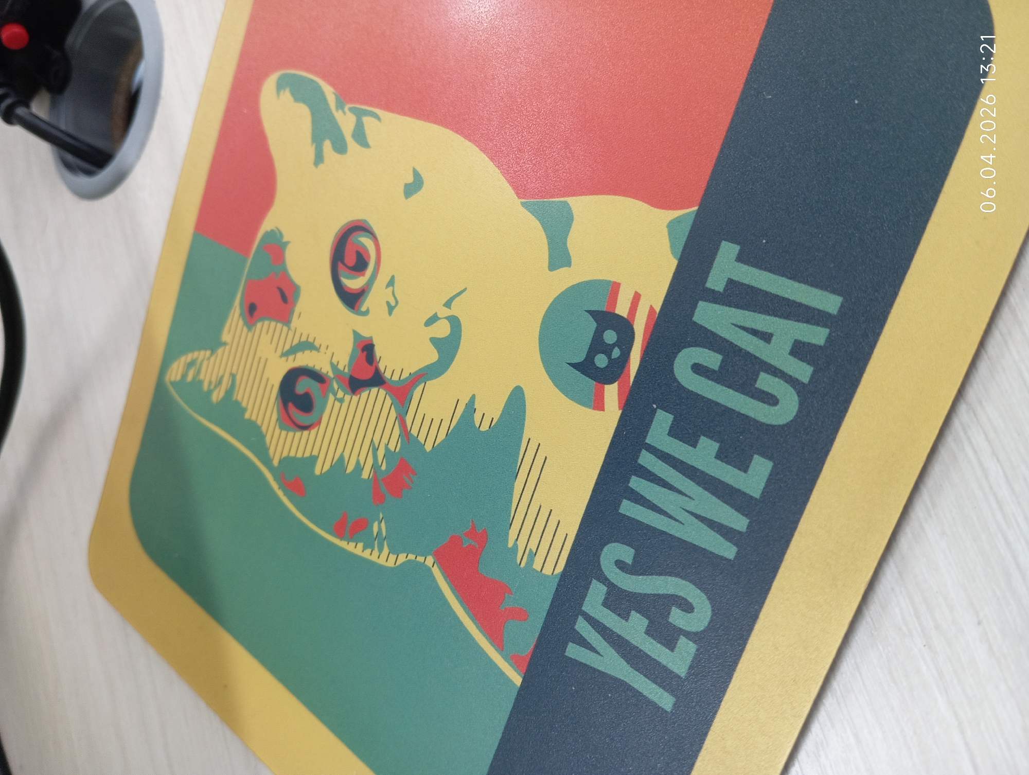 The image shows a mousepad with a stylized illustration of a cat. The illustration is divided into three color blocks: red, yellow, and green. The cat illustration is mostly yellow with red and green accents. A dark blue banner is placed across the illustration, and it has the words "YES WE CAT" written on it in green. The date "06.04.2026 13:21" is printed vertically in the top right corner of the mousepad. A partially visible power outlet with a red button is in the upper-left corner of the image. The mousepad appears to be on a desk. The location is not discernible from the image.