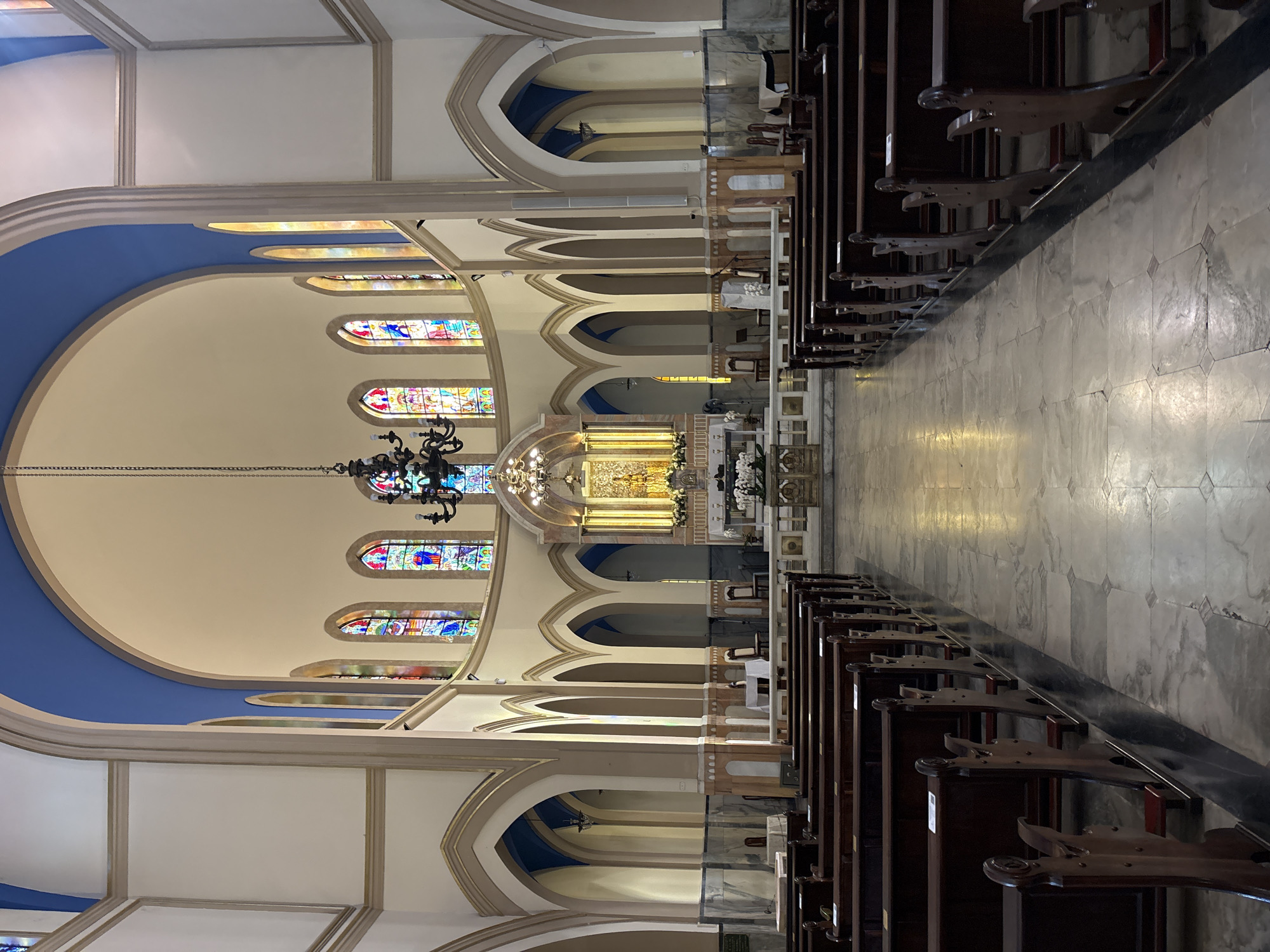 The image shows the interior of the Catedral da Sé in São Paulo, Brazil. The vast, open space of the cathedral is visible, with rows of dark wooden pews stretching down the central aisle. The floor is made of polished marble with a geometric pattern. The walls and vaulted ceiling are painted in neutral tones, with the ceiling featuring a striking blue expanse. Stained-glass windows, depicting colorful religious scenes, line the upper walls of the nave. A large, ornate chandelier hangs from the ceiling, and a golden altar is visible at the far end of the cathedral, adorned with flowers. The scene is empty of people, creating a sense of quiet solemnity.
