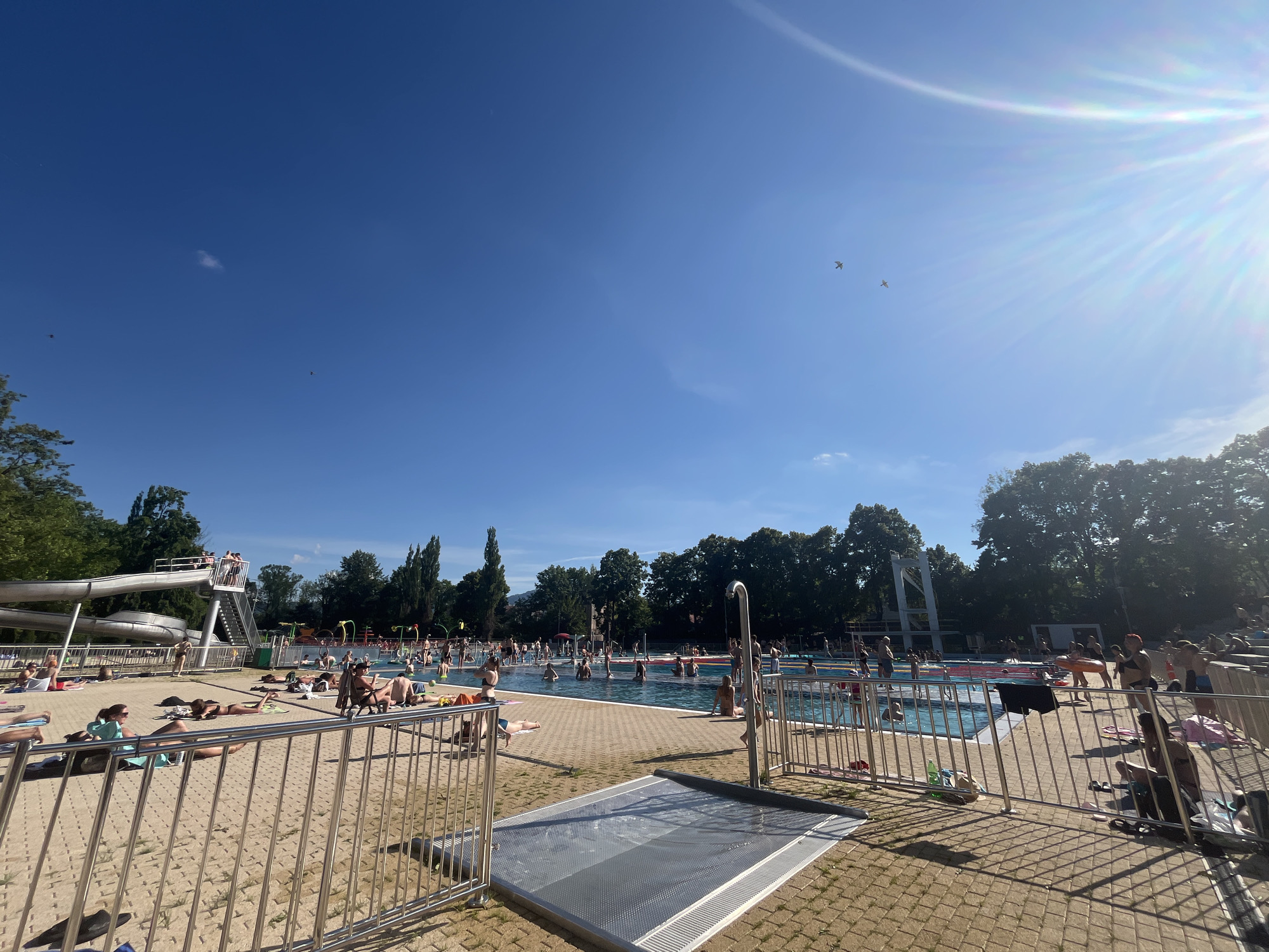 The image shows a crowded outdoor swimming pool on a sunny day. In the foreground, a metal fence separates the viewer from a paved area with people sunbathing and relaxing on towels and chairs. Beyond the fence, the main pool area is visible, filled with many people swimming, wading, and standing in the water. There are water slides and other pool amenities in the background, along with trees and buildings bordering the pool. The sky is clear and blue, with the sun prominently visible in the upper right corner, causing a lens flare. The overall scene depicts a lively and popular recreational area.