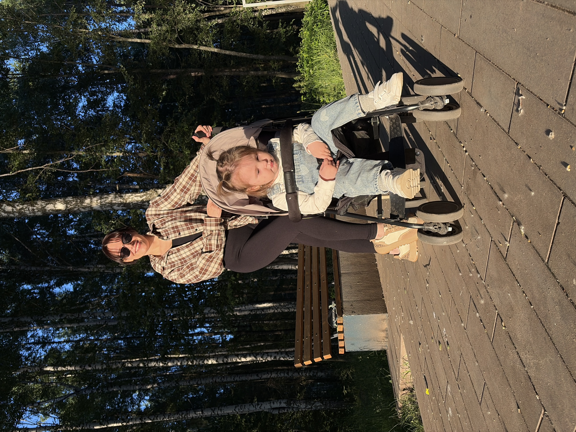 In the city of Chaikovsky, Russia, a woman with dark hair and sunglasses stands behind a stroller on a paved pathway. She is wearing a brown and white checkered shirt over a black top and dark leggings. The woman is smiling and looking at the camera. Seated in the stroller is a young child with blonde hair styled in pigtails. The child is wearing a white long-sleeved shirt and blue denim overalls, with grey socks and white sneakers. The child is looking down and appears somewhat pensive. The background features a dense forest of birch trees with dark green foliage, and a wooden park bench is visible to the left of the pathway. The scene is illuminated by the warm, late afternoon sun, casting long shadows on the ground.