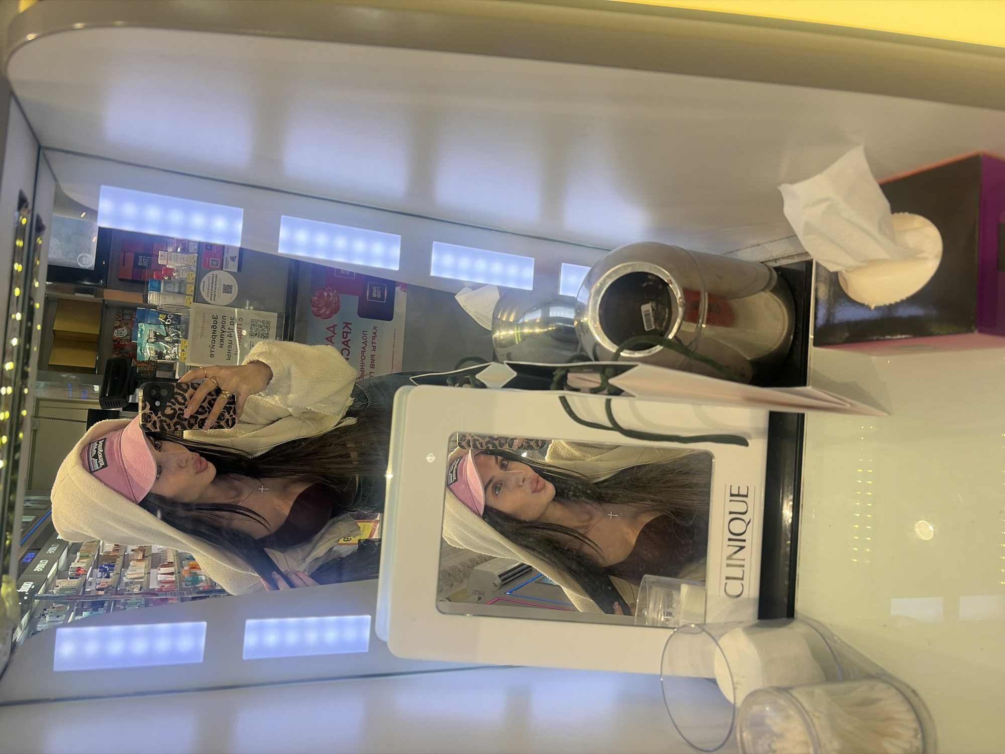 A young woman is taking a selfie in a brightly lit store, likely a beauty or cosmetic shop, given the shelves of products visible in the reflection of the mirror she's using. She is wearing a light-colored hooded jacket and a pink baseball cap. In the mirror, she is reflected looking into her phone, which has a leopard-print case. A small cross necklace is visible around her neck. The reflection also shows a display of Clinique products, including a prominent "CLINIQUE" logo. The store appears to be indoors with fluorescent lighting, and the time of day is indeterminate due to the artificial illumination. The overall mood seems casual and self-aware. The visible text on the Clinique display is "CLINIQUE". Other text in Cyrillic, including "АД" and other words related to sales or promotions, is also present on packaging behind her.