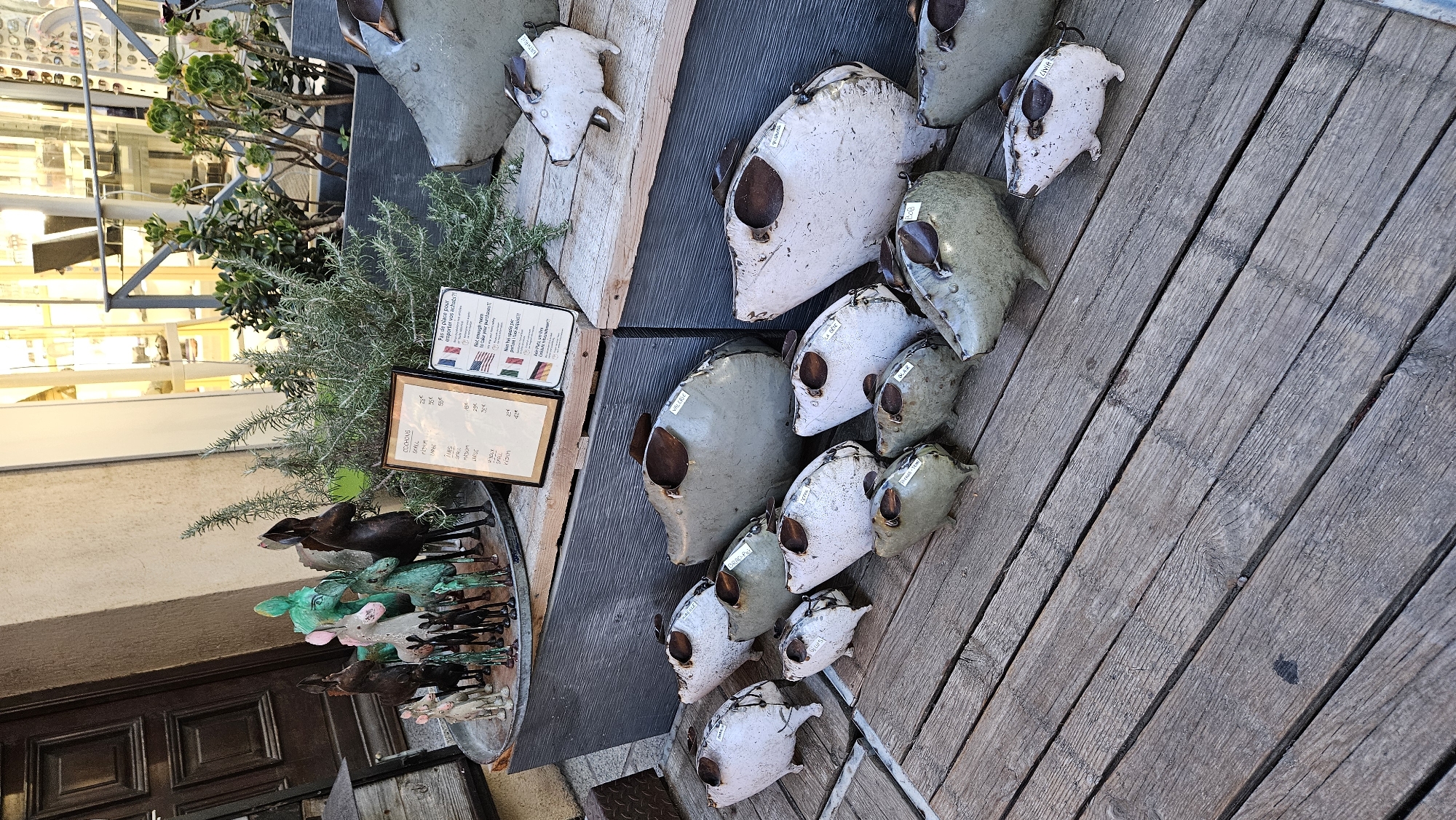 This image captures an outdoor display of decorative metal pigs and donkey figurines in L'Île-Rousse, France. The subjects are arranged on a weathered wooden platform in front of a building with a dark wooden door and large windows showcasing merchandise. Several ceramic planters with green succulents and rosemary are positioned behind the figurines.
The metal pigs are of various sizes and colors, predominantly white, gray, and a muted green. Some have small labels attached, indicating prices or names. The donkey figurines, in shades of green and dark brown, are clustered on a circular metal tray in the background. Two framed signs are also visible; one appears to be a price list, and the other features the flags of France, the USA, and Germany with multilingual text. The overall scene suggests a retail display, likely for garden ornaments or souvenirs, in a quaint European setting.