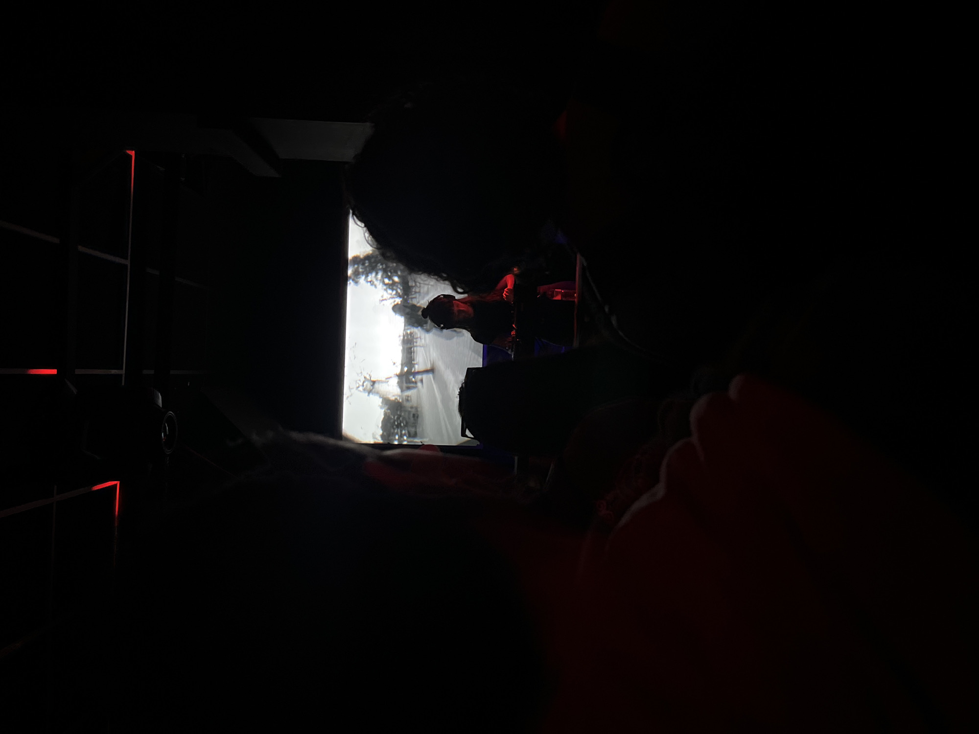A person with dark hair watches a brightly lit television in a dark room. Red light emanates from an unknown source on the left side of the frame. The screen displays an image that is predominantly white and blurry, appearing to show a cityscape. The person is facing toward the television. Part of the person's foot is visible in the lower right. The location in the city of Barcelona, Spain is not identifiable in the image.
