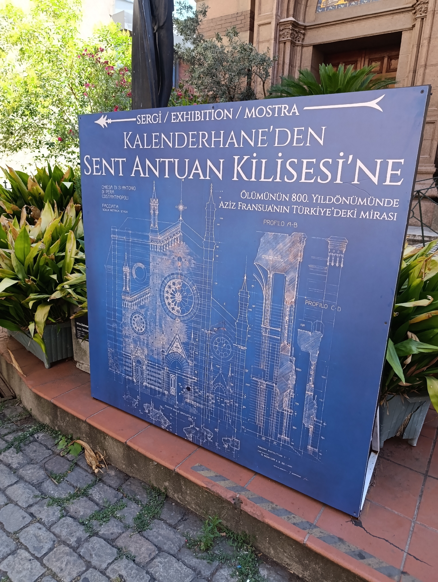 The image shows a large blue sign with white architectural drawings and text displayed outdoors, likely in the city of Beyoğlu, Turkey. The sign is mounted on a stand and is positioned on a paved area next to a brick ledge. Lush green plants are visible in planters to the left and right of the sign. The text on the sign appears to be in both Italian and Turkish and references an exhibition about the "Church of Saint Anthony of Padua" (Chiesa di S. Antonio di Pera) in Constantinople. The architectural drawings depict detailed blueprints of the church. The environment suggests an urban setting with historical architecture, as suggested by the brickwork and the presence of a building facade in the background. No people are visible in the image.