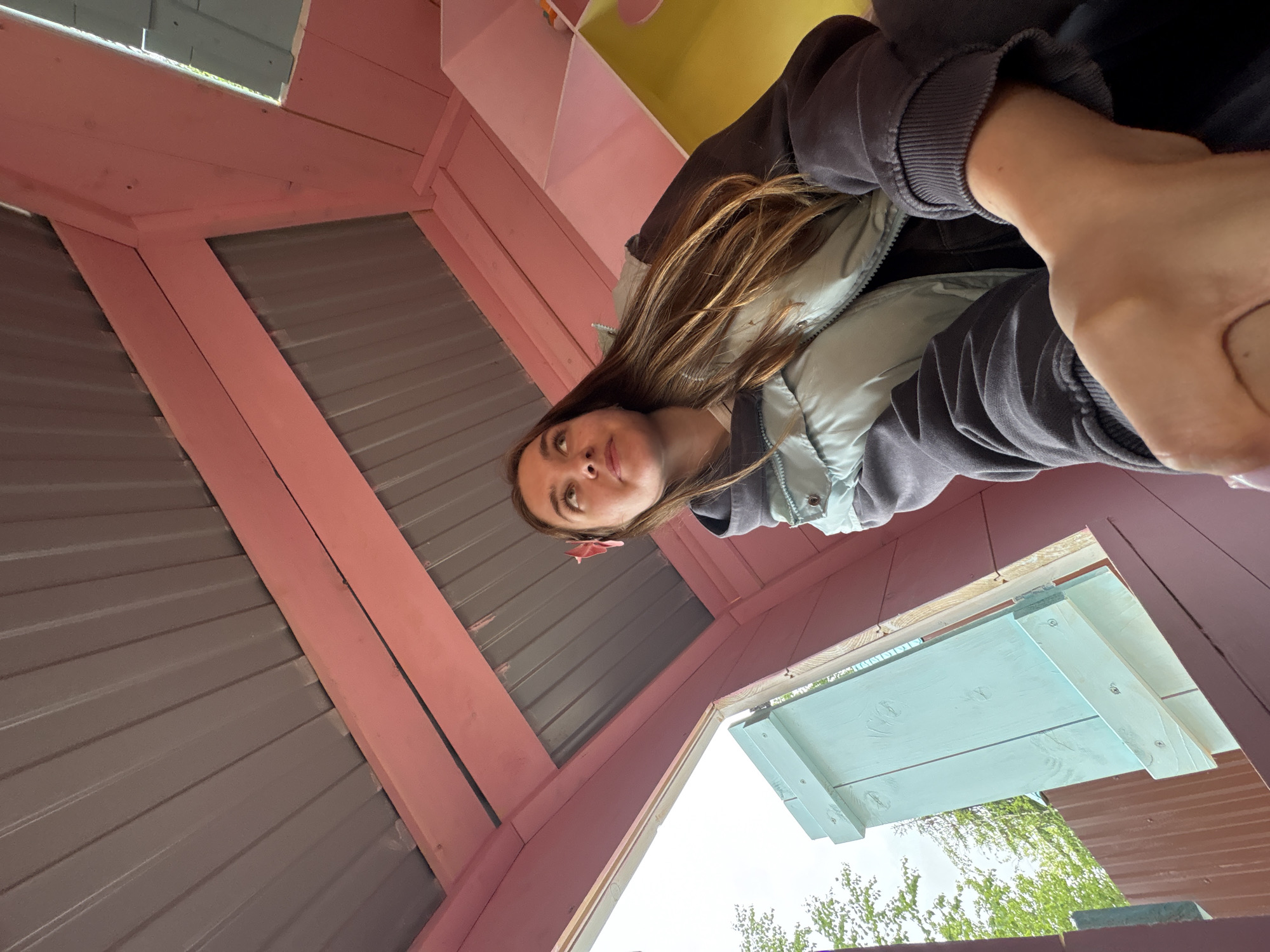 A low-angle shot depicts a woman inside a structure with pink and grey construction. The woman is wearing a grey jacket over a light blue puffer jacket and has long brown hair. She looks upwards, smiling faintly. The building interior has a combination of pink wooden beams and grey corrugated metal. A window shows greenery outside. The location is Perm, Russia.