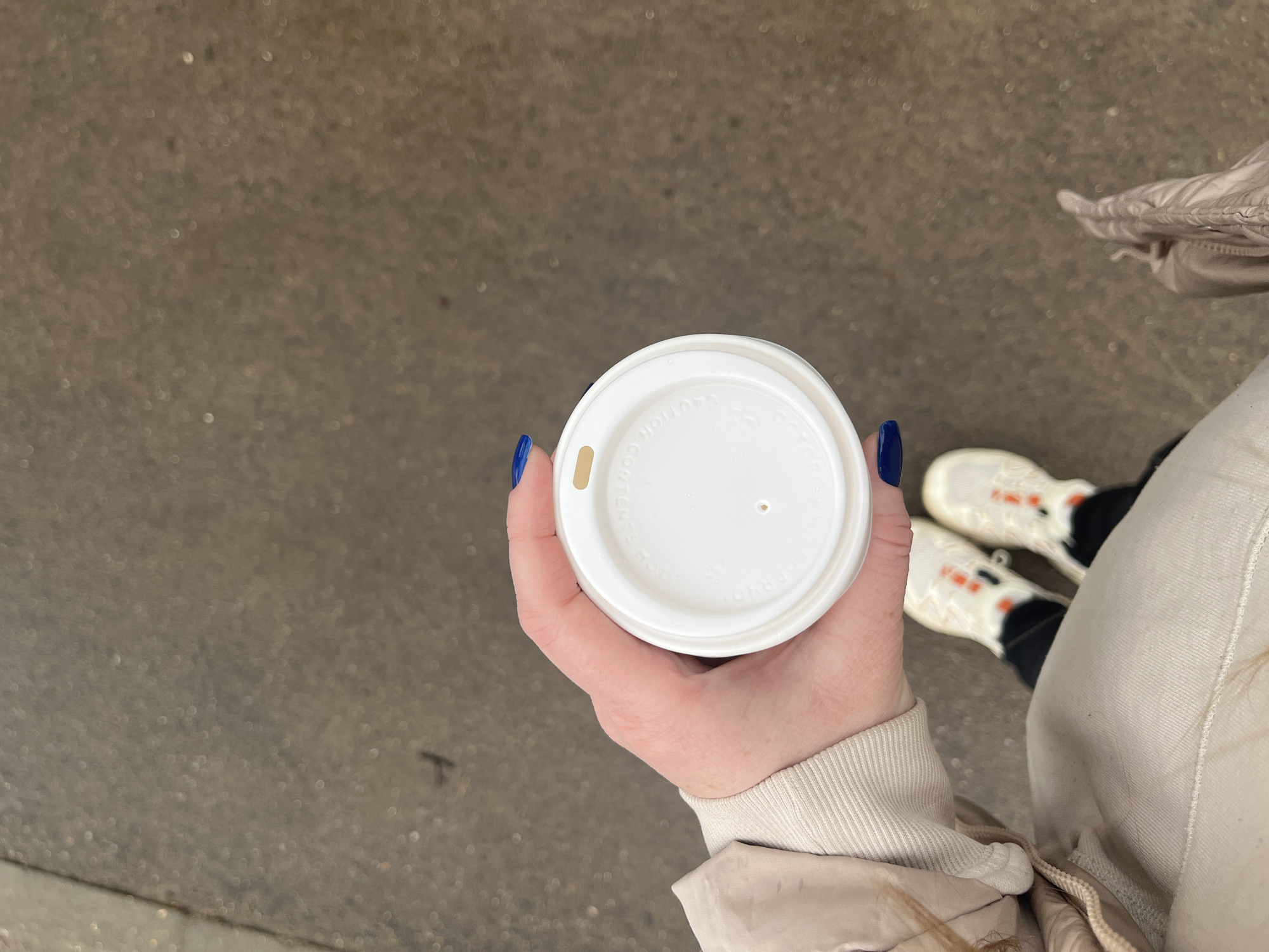The image shows a person holding a white disposable coffee cup with a white lid, which has a small hole in the center and a sliding vent. The person is wearing a light-colored jacket and white sneakers with black and orange accents. Their fingernails are painted dark blue. The background is a grey concrete surface, suggesting an urban environment. There is no evidence in the image that directly identifies the location as Луговой, Russia.