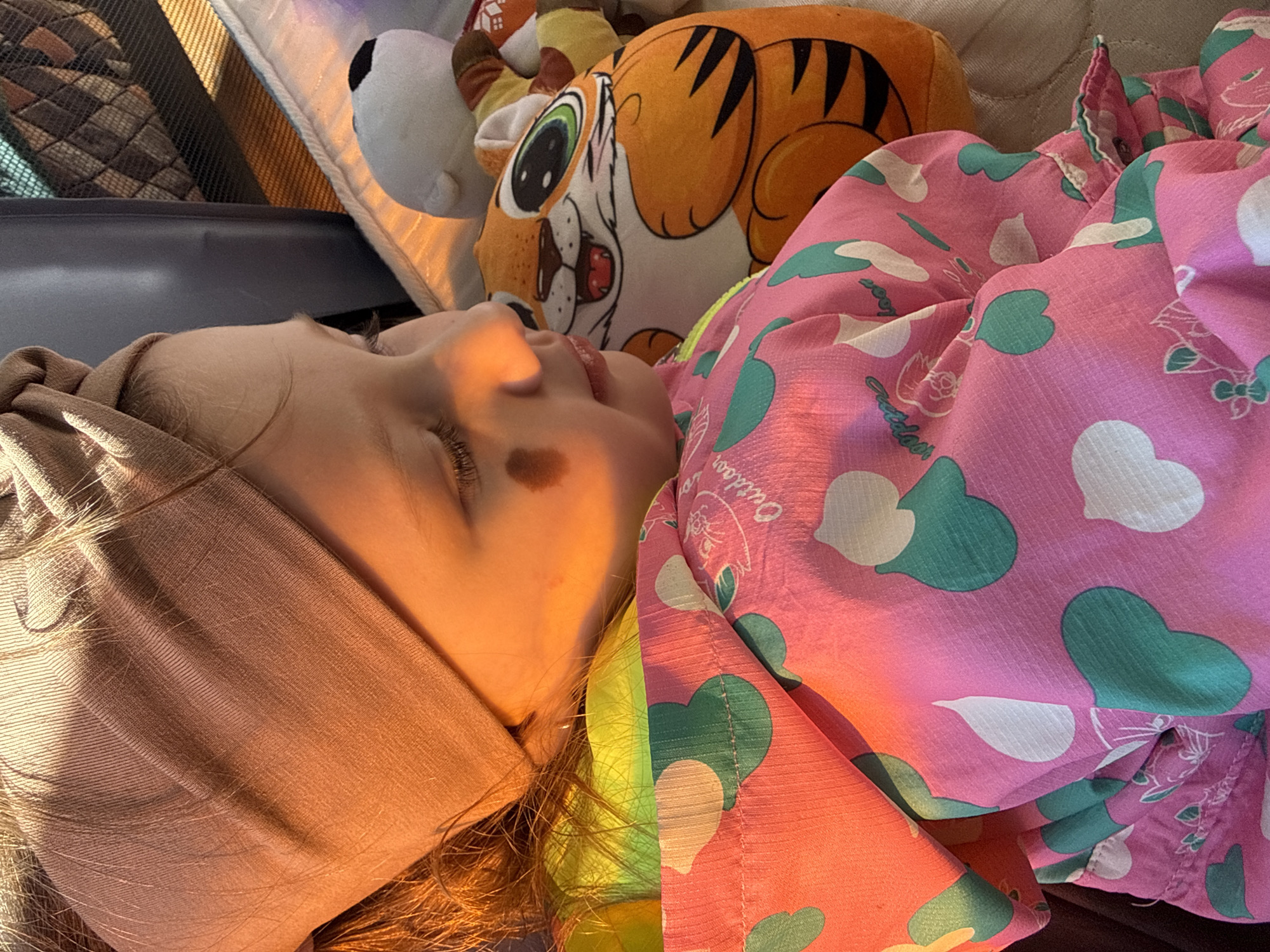 A child with a tan head wrap is sleeping on their side next to a tiger plush toy. They are wearing a pink and green heart-patterned shirt. The location is likely a car or bus. The specific city, Кипчак, Russia, cannot be verified from the image alone.