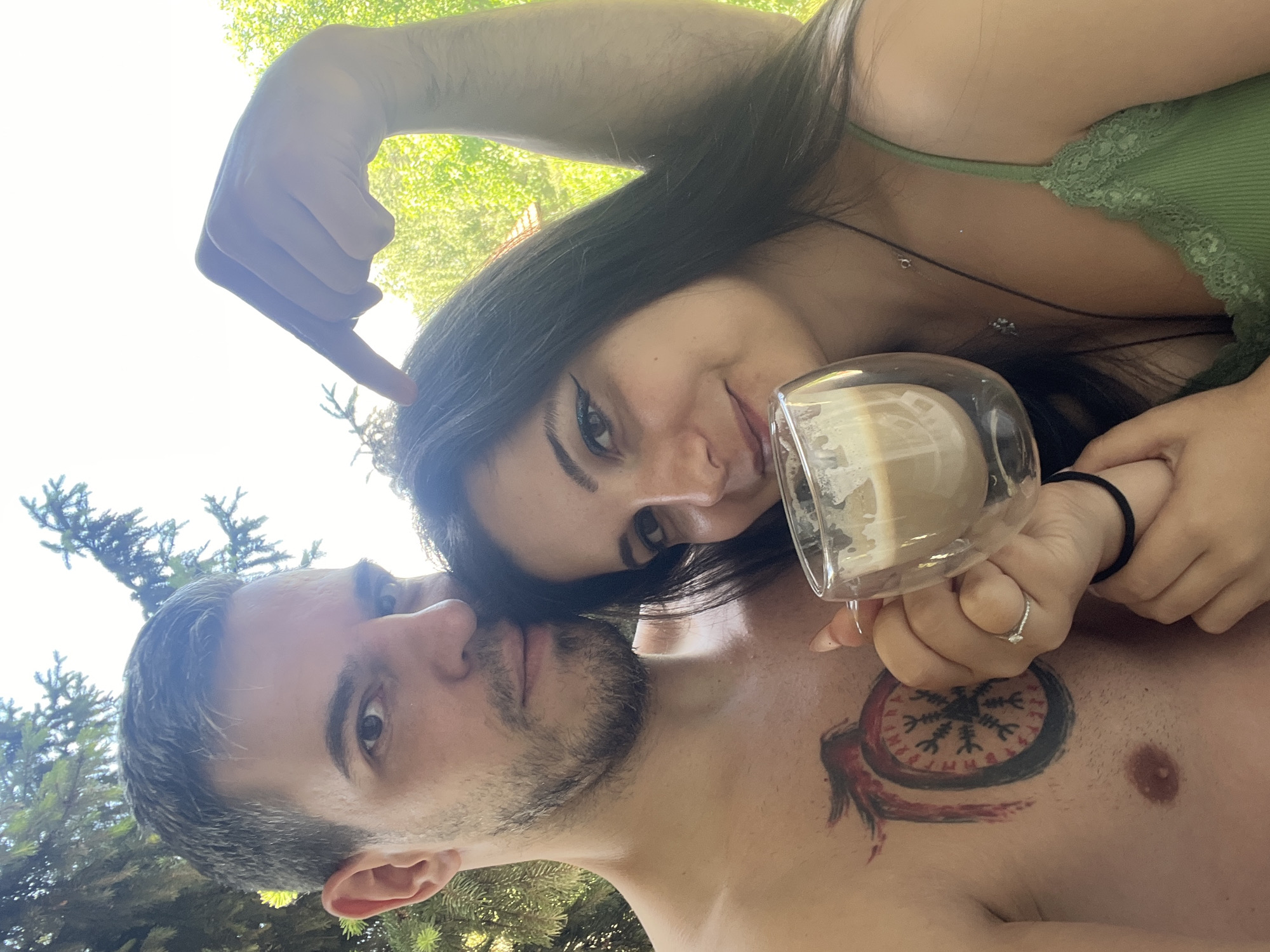 A man and a woman are posing for a selfie outdoors in Sofia, Bulgaria. The man is shirtless, revealing a large tattoo on his chest. The woman is holding a clear mug of coffee and smiling at the camera. They are positioned close together, with the man's arm around the woman's shoulders and his finger pointing towards her head. The background consists of lush green trees and foliage, suggesting a park or garden setting. The sunlight indicates it is daytime.
